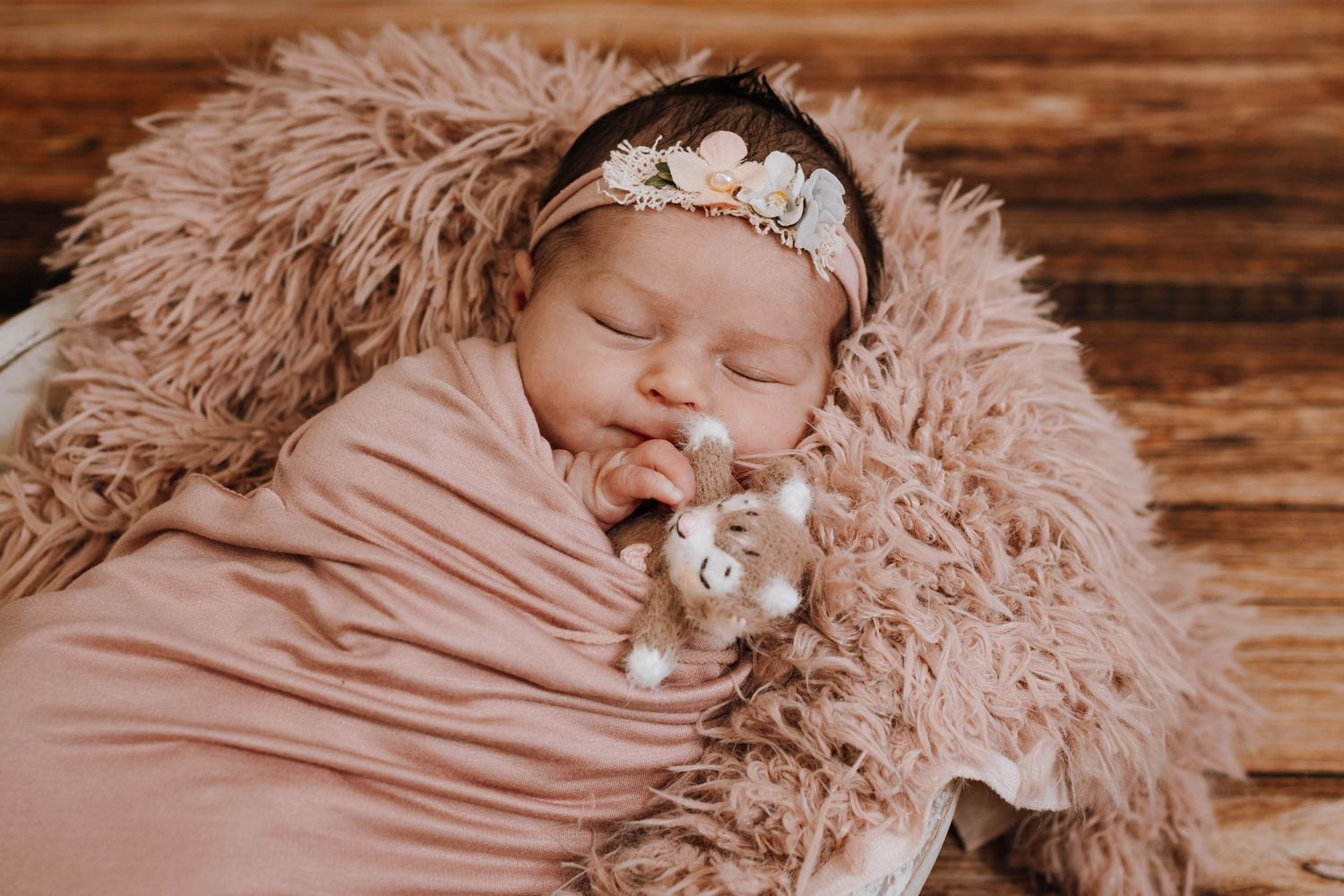 Sleeping newborn wrapped in pink swaddle with delicate headband on fluffy pink backdrop.