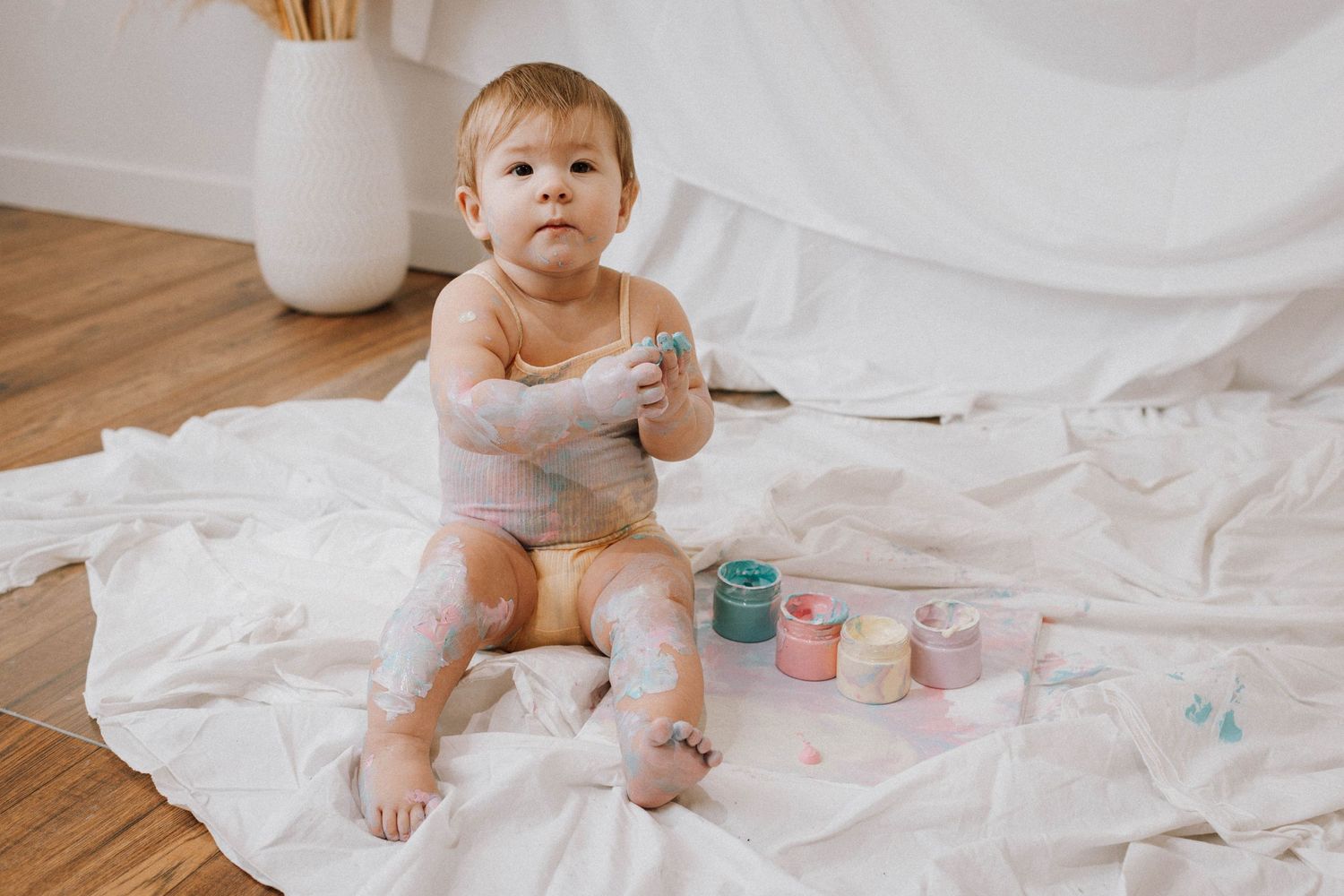 Baby in light colored outfit sitting on white blanket with colorful paint containers.
