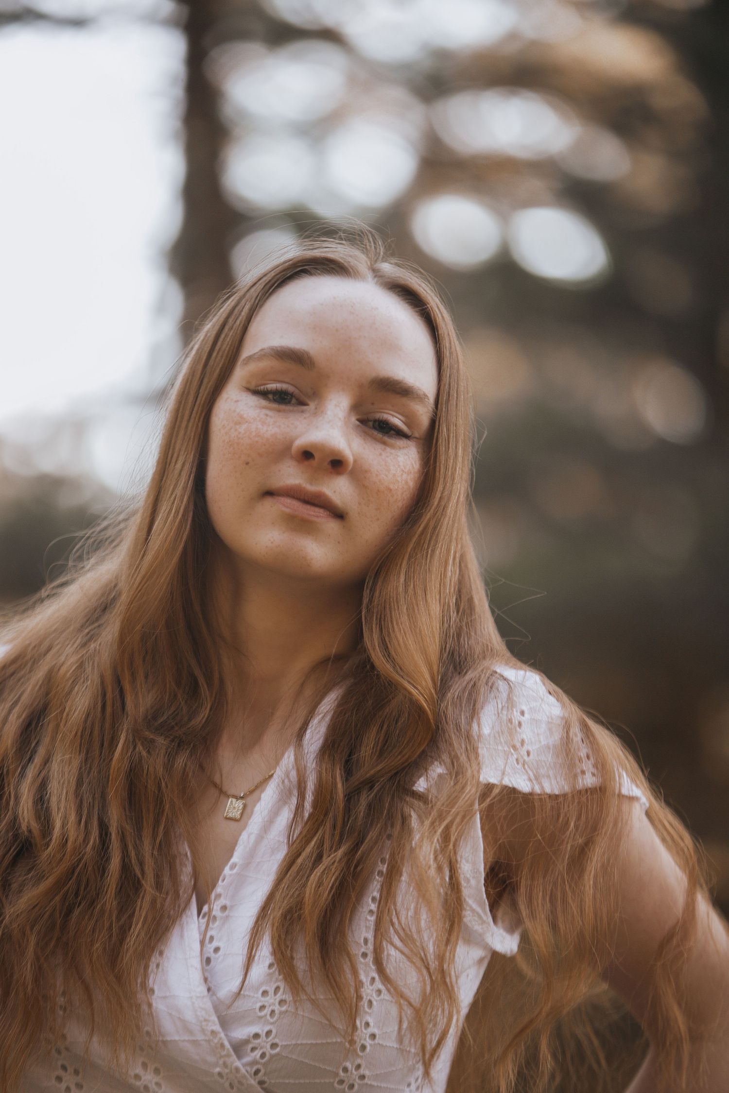 Young woman in white lace dress poses outdoors during golden hour with soft bokeh background.