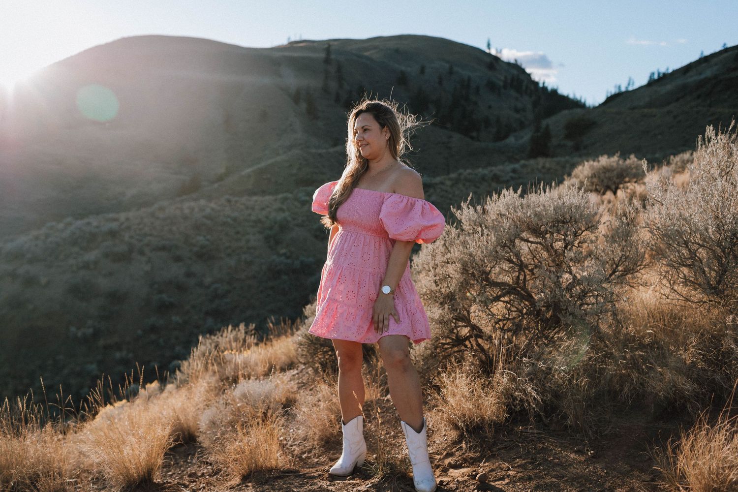 A person in a pink dress poses on a mountain trail with sun flare in the background.