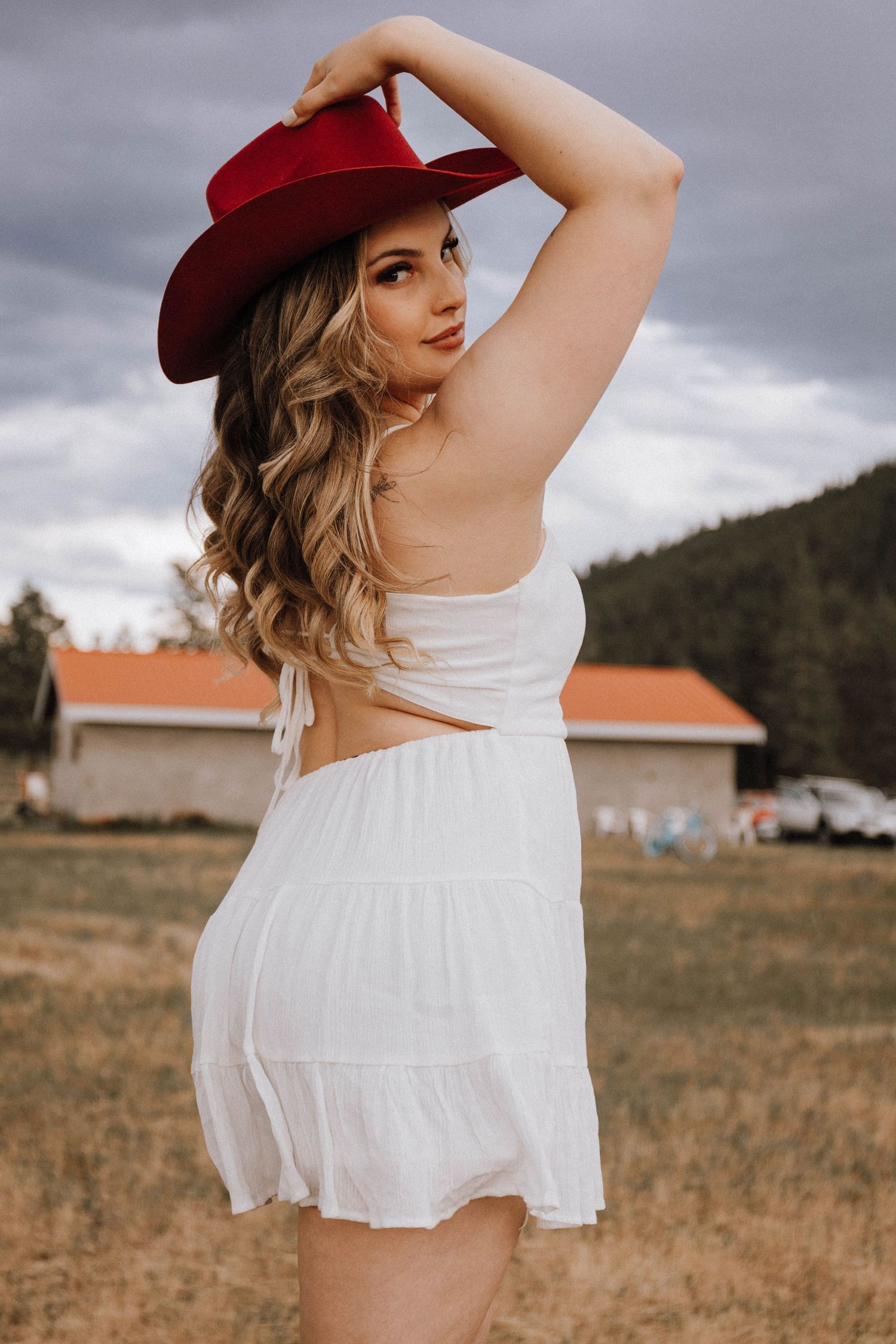 A person in a white sundress and red cowboy hat poses outdoors against a cloudy sky and rural backdrop.
