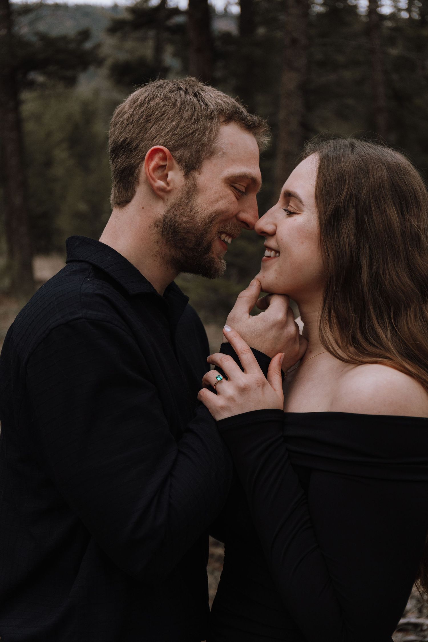 A couple in matching black outfits share a romantic almost-kiss moment outdoors surrounded by pine trees.