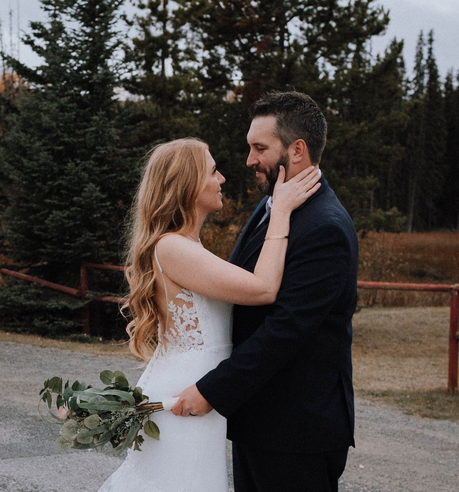 A romantic wedding couple embraces on a mountain path surrounded by evergreen trees.
