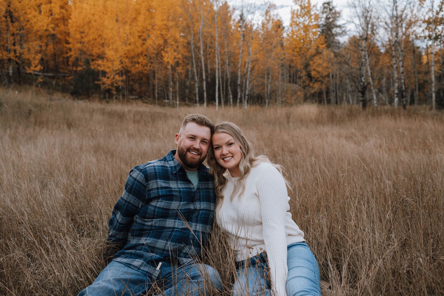 Couple shares intimate moment in tall grass with golden autumn trees behind them.