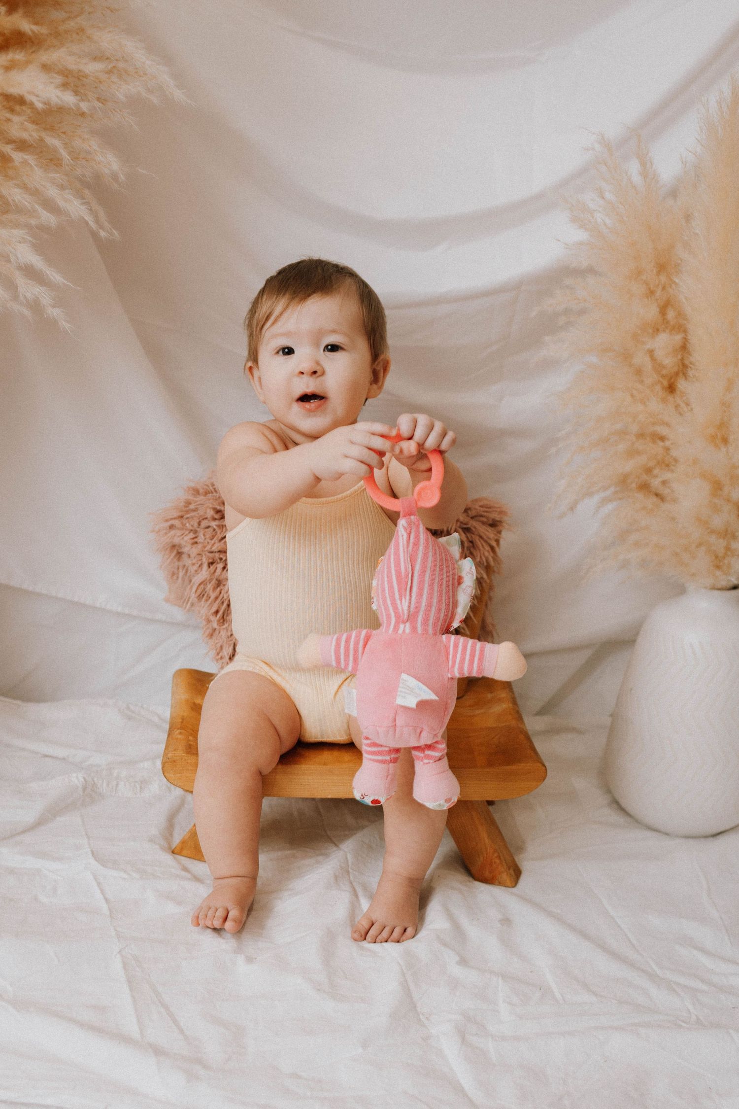 Baby in beige romper sits on wooden stool holding pink plush toy with pampas grass decor in background.