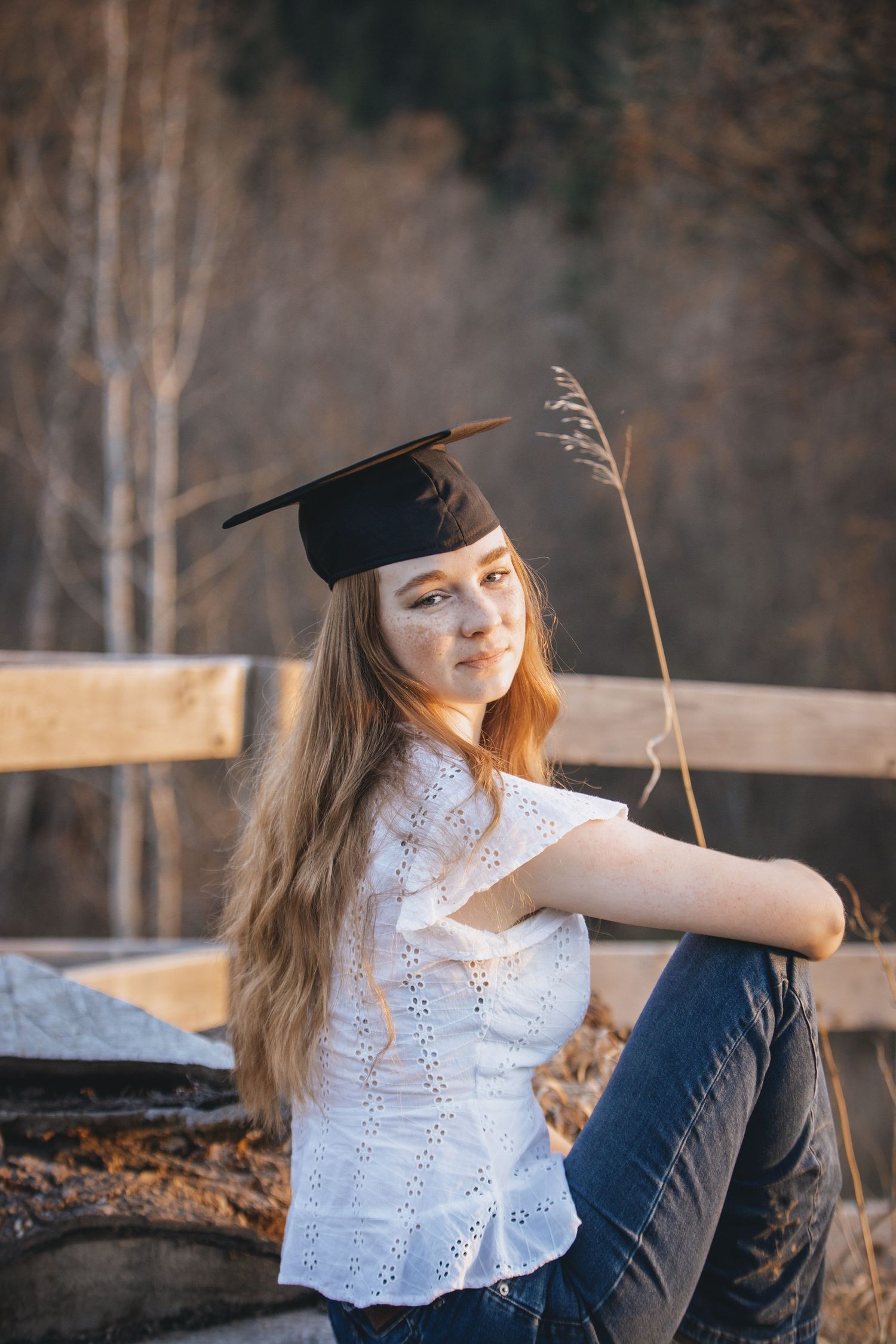 Graduate in cap and white eyelet top sits casually on wooden fence in autumn forest.