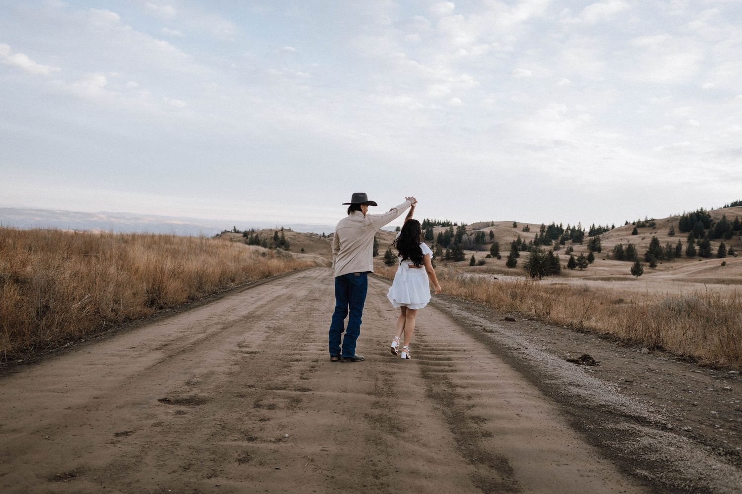A couple walking hand in hand down a dirt road through golden fields on a cloudy day.