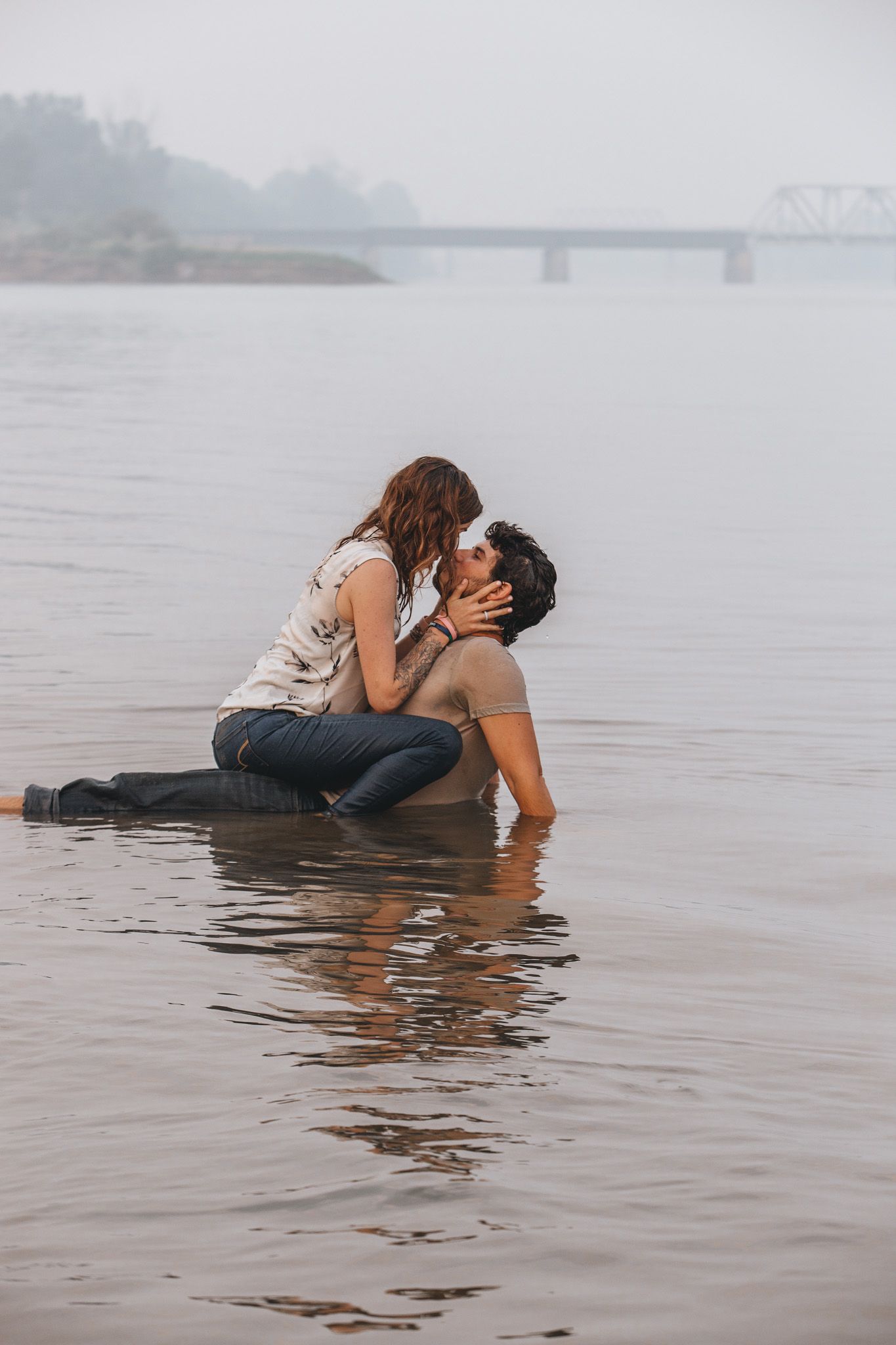 A couple shares an intimate moment while lying in shallow water at the edge of a misty lake.
