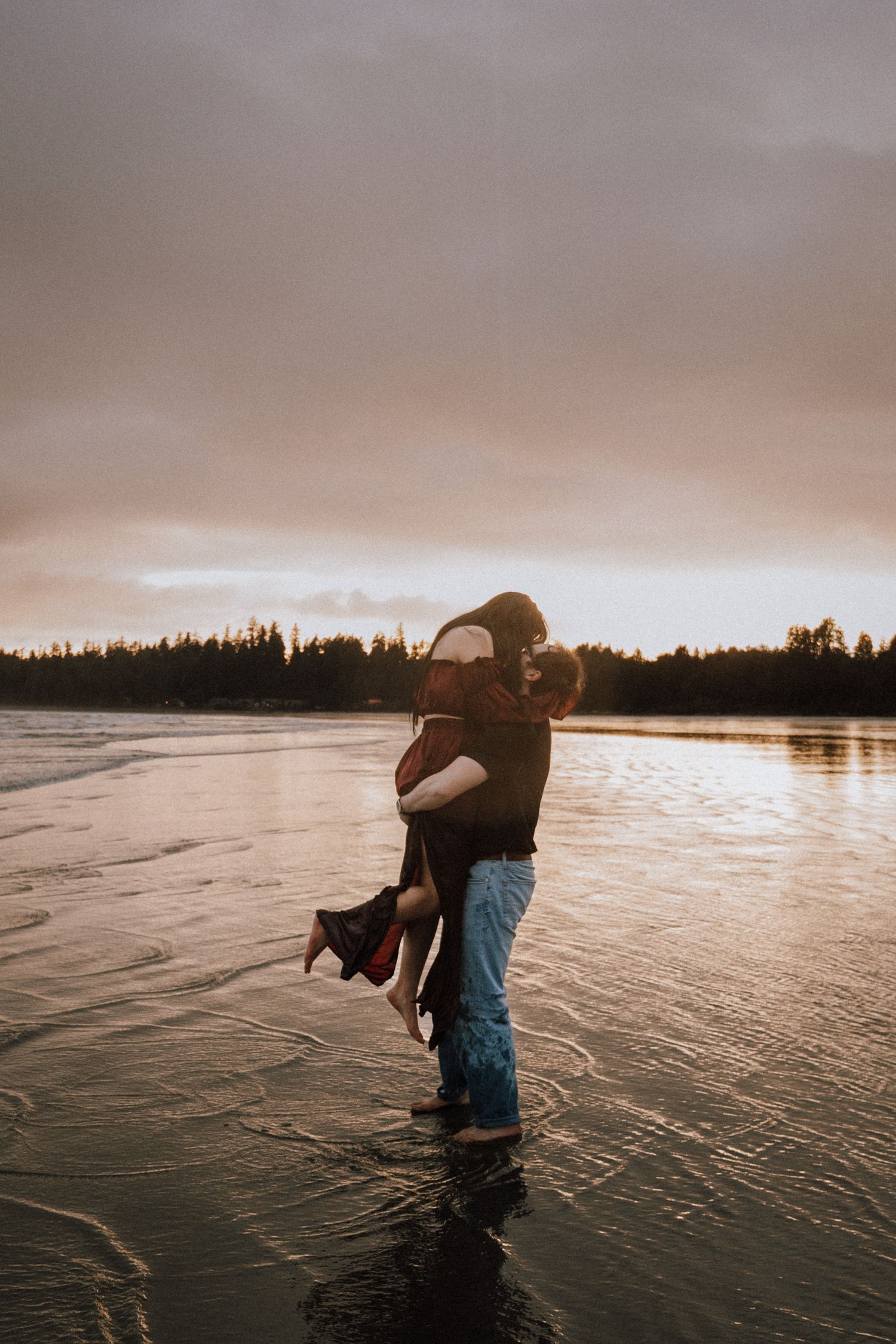 Couple embracing on a misty beach at sunset, man lifting woman near shoreline with trees in background.