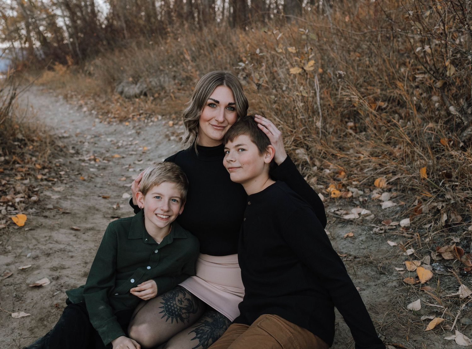 A woman and two children sitting together on a nature path surrounded by autumn foliage.