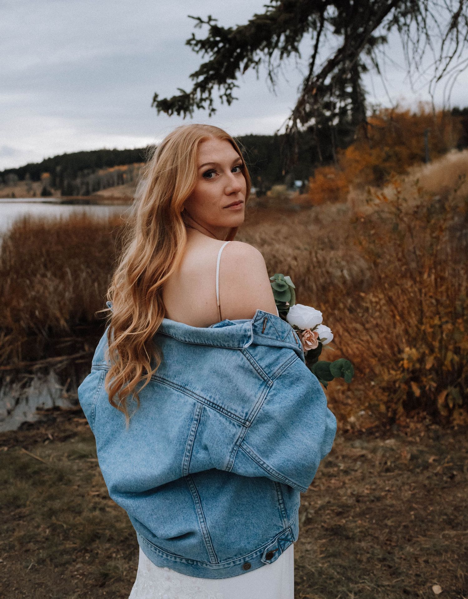 A woman in a white dress and denim jacket poses outdoors against an autumn landscape with mountains and a lake.