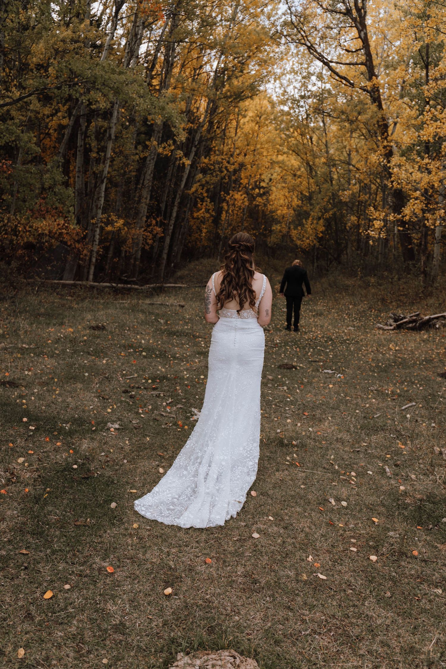 A bride in a lace gown stands in a forest with autumn foliage.