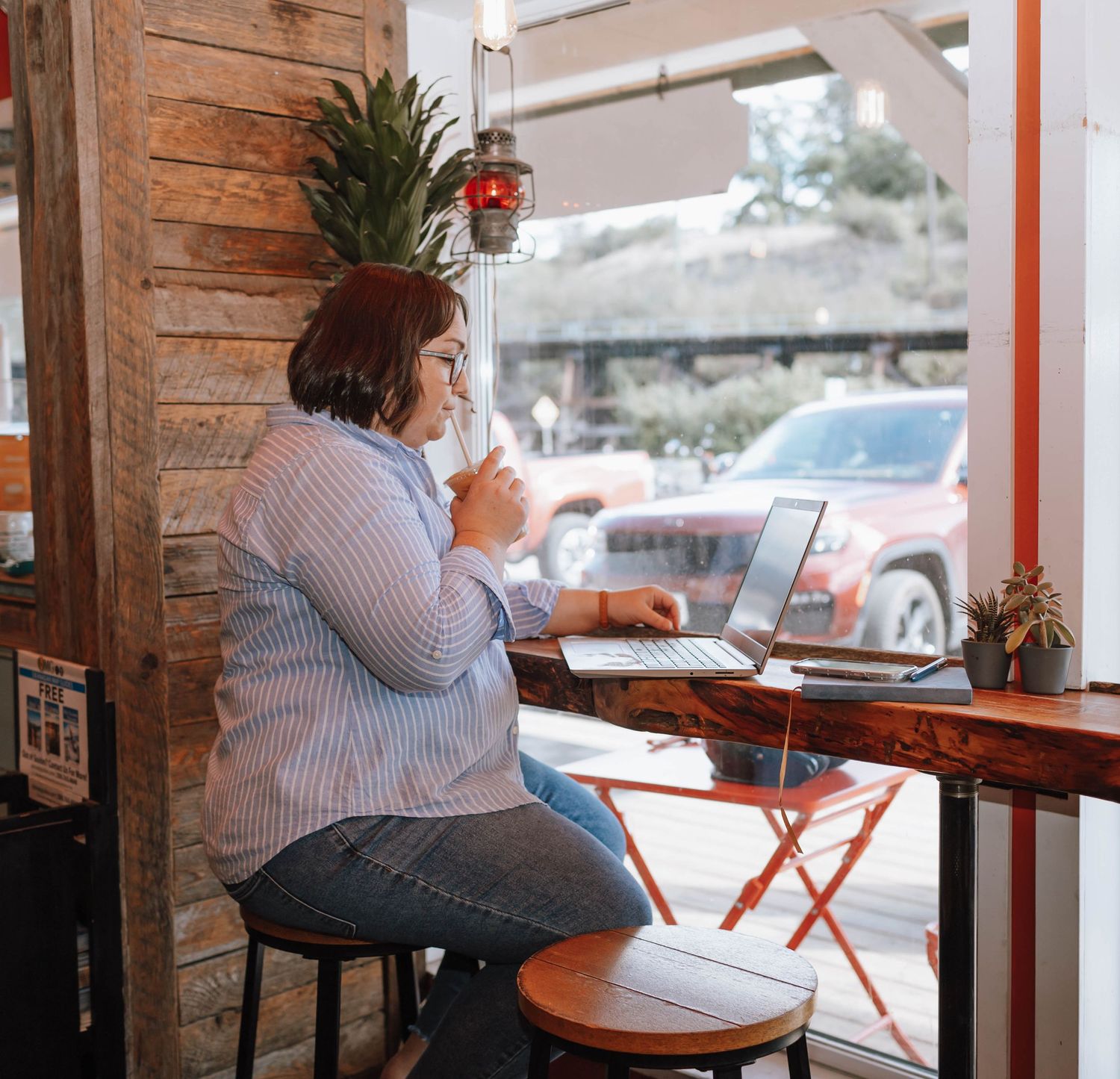 Person enjoying beverage while working on laptop at rustic cafe window seat.