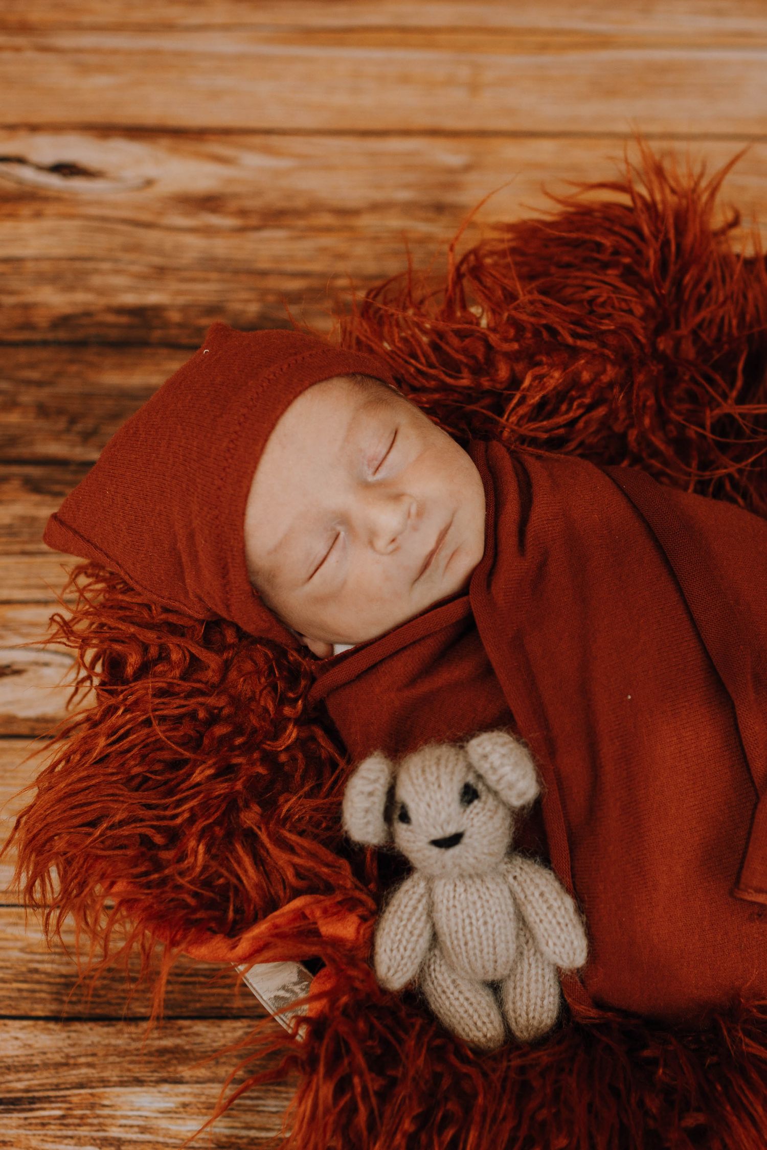 A sleeping baby swaddled in deep red fabric lies on a fluffy burgundy rug while holding a small knitted teddy bear toy.