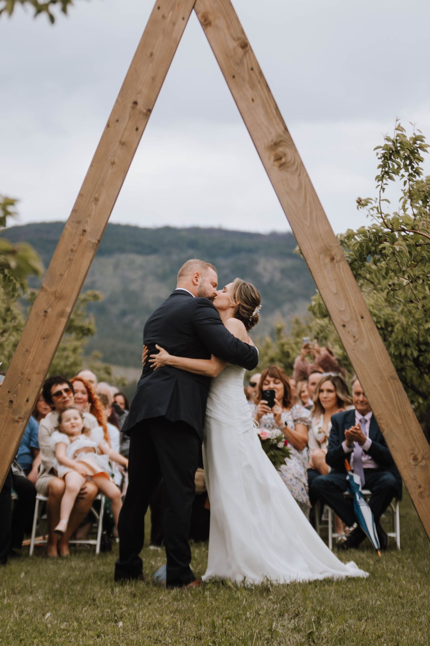 Wedding ceremony kiss under wooden triangle arch surrounded by guests on a grassy hillside with mountain backdrop.