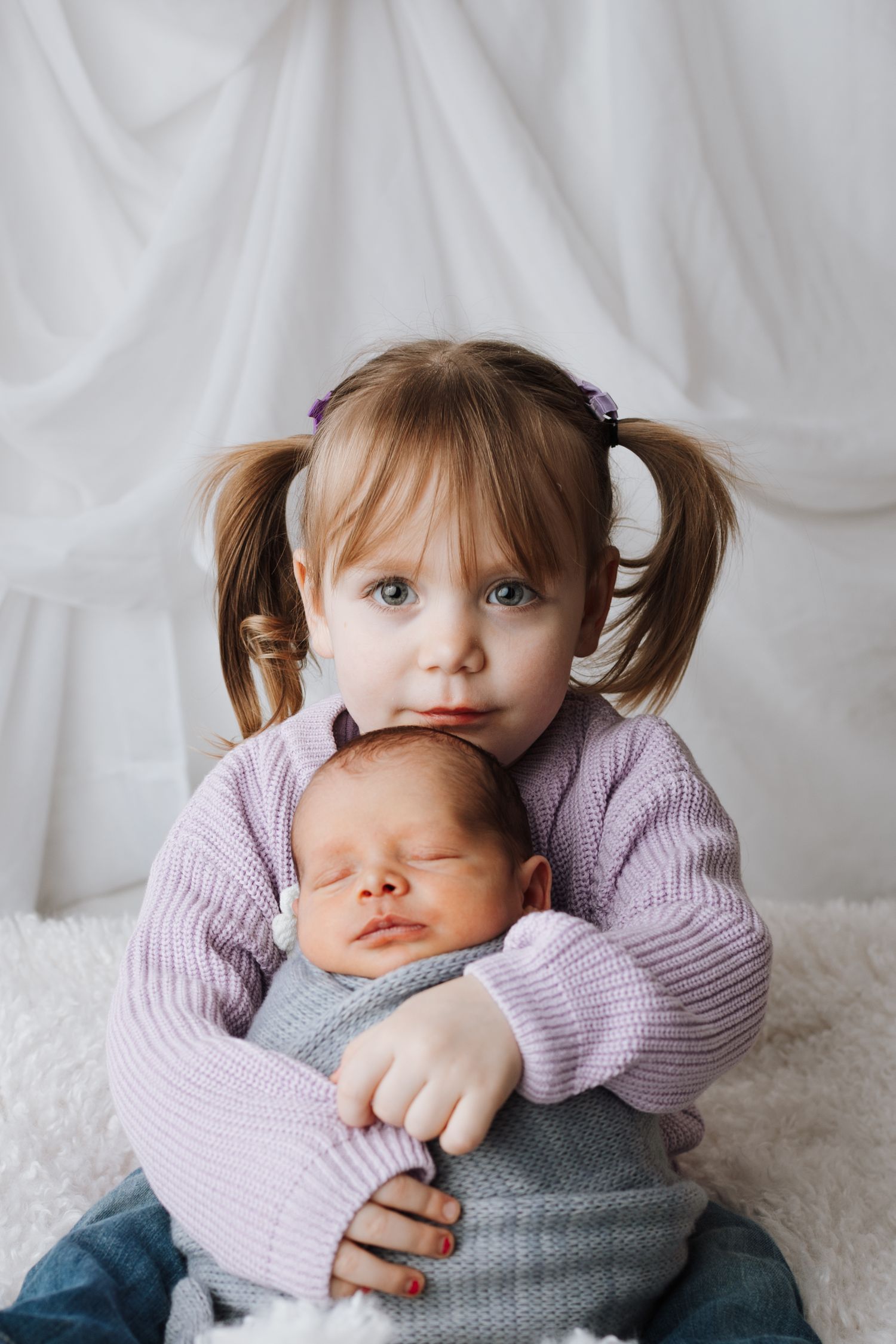 Young girl with pigtails tenderly holding sleeping newborn baby sibling, both dressed in soft pastel colors.