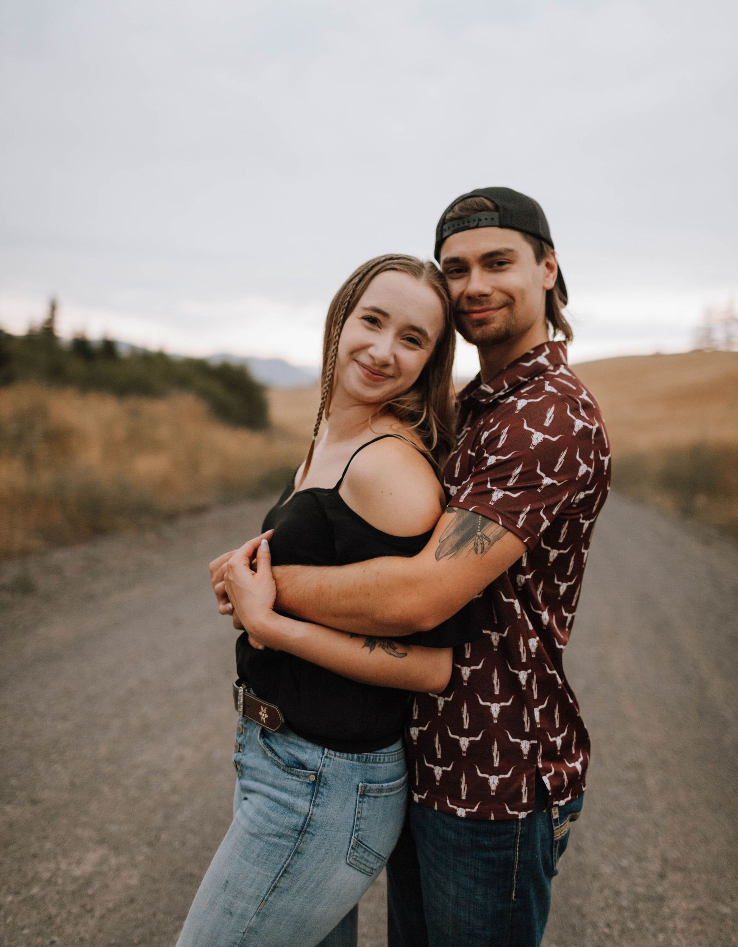 Couple embraces on a dirt road with mountains and cloudy skies in the background.