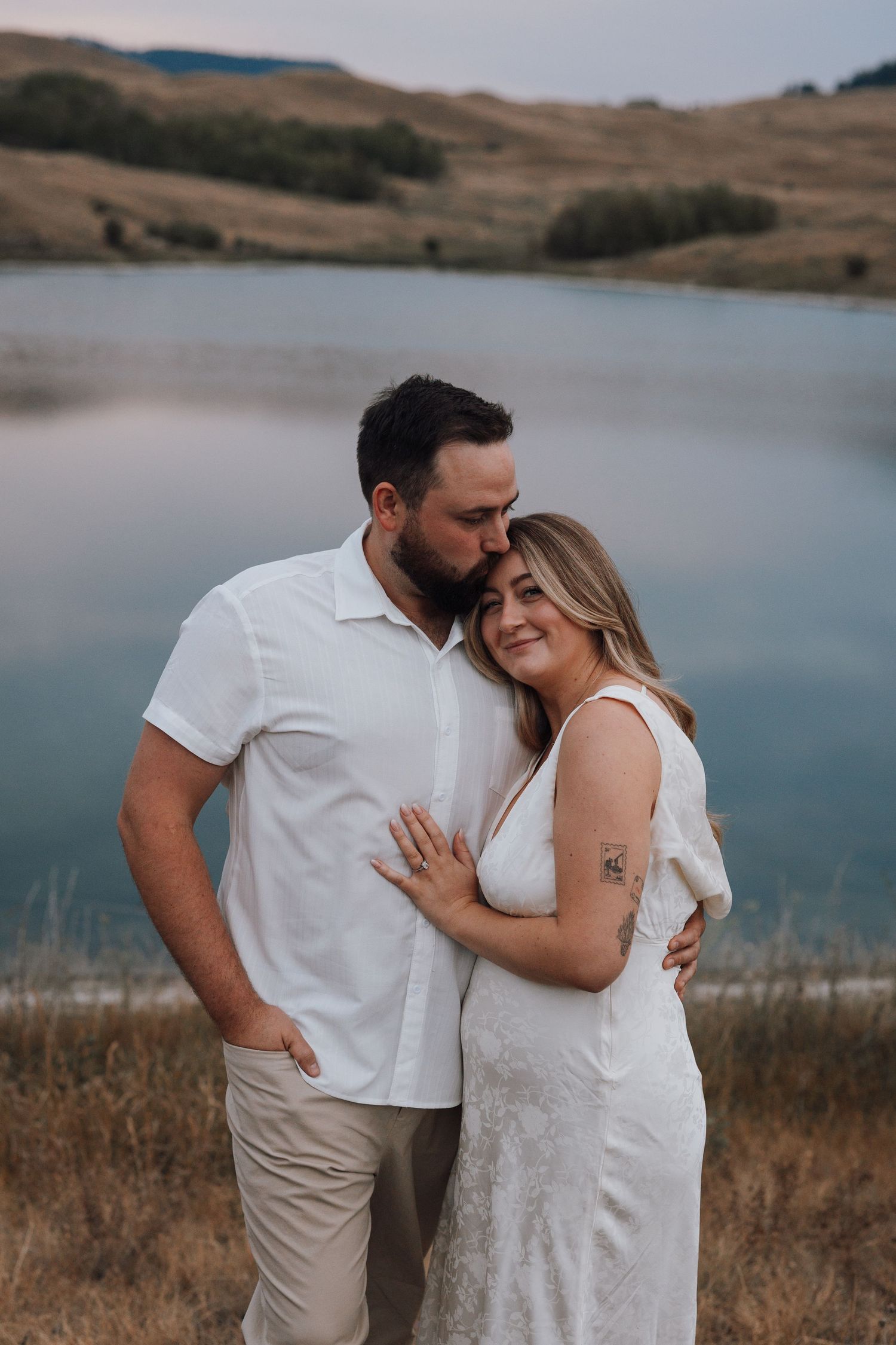 Couple in white outfits embracing near a serene lake with rolling hills, man kissing woman's head during engagement photo.
