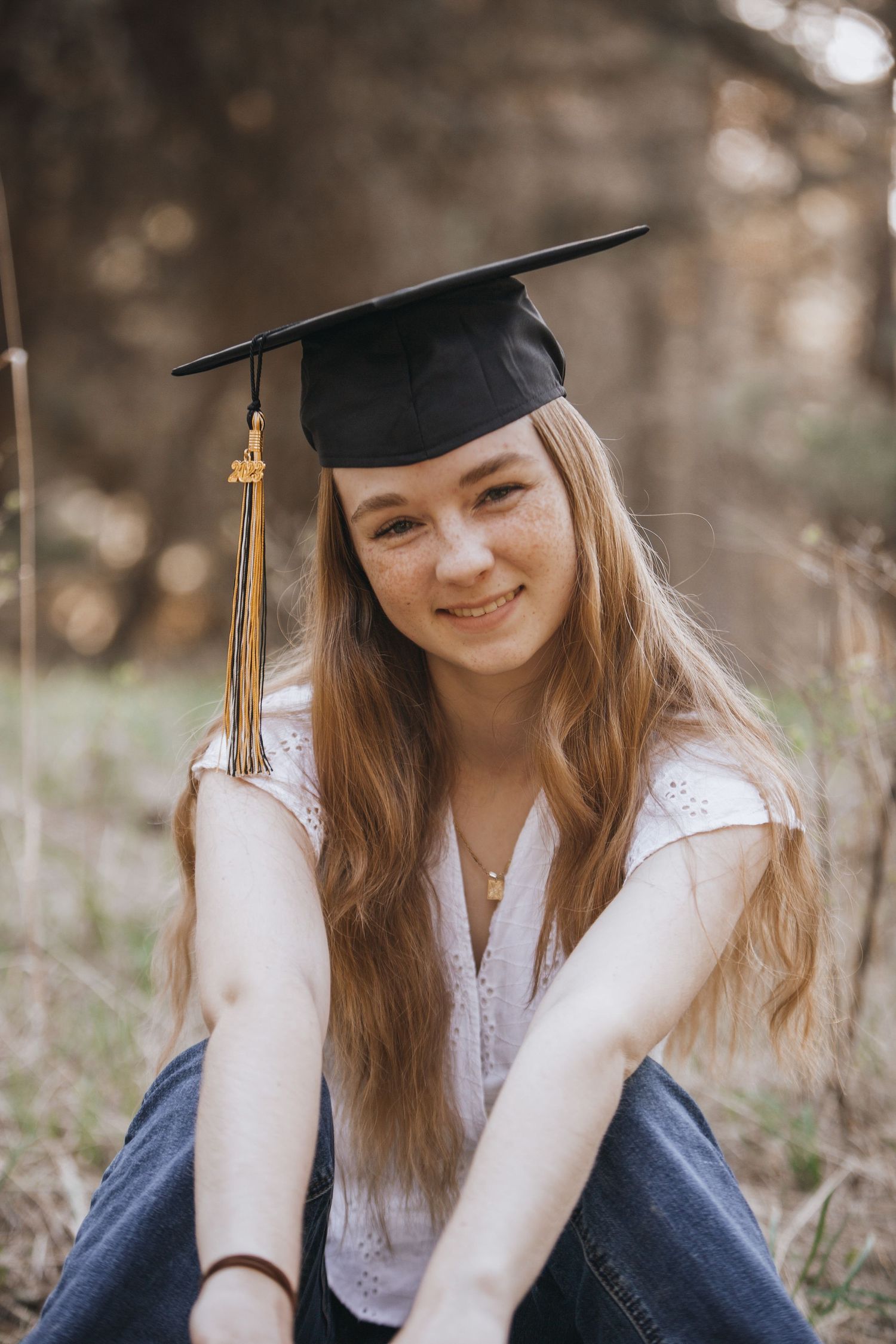 Graduate in cap and gown sits outdoors in casual graduation portrait.