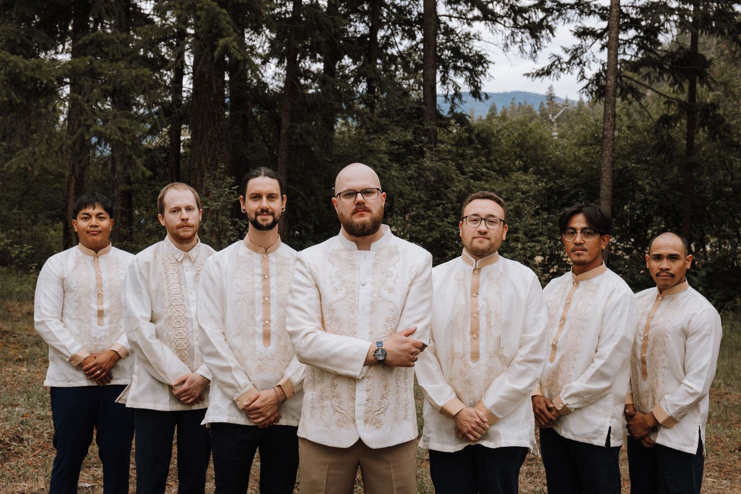 A group in matching white button-down shirts pose together in a forest setting with mountains visible in the background.