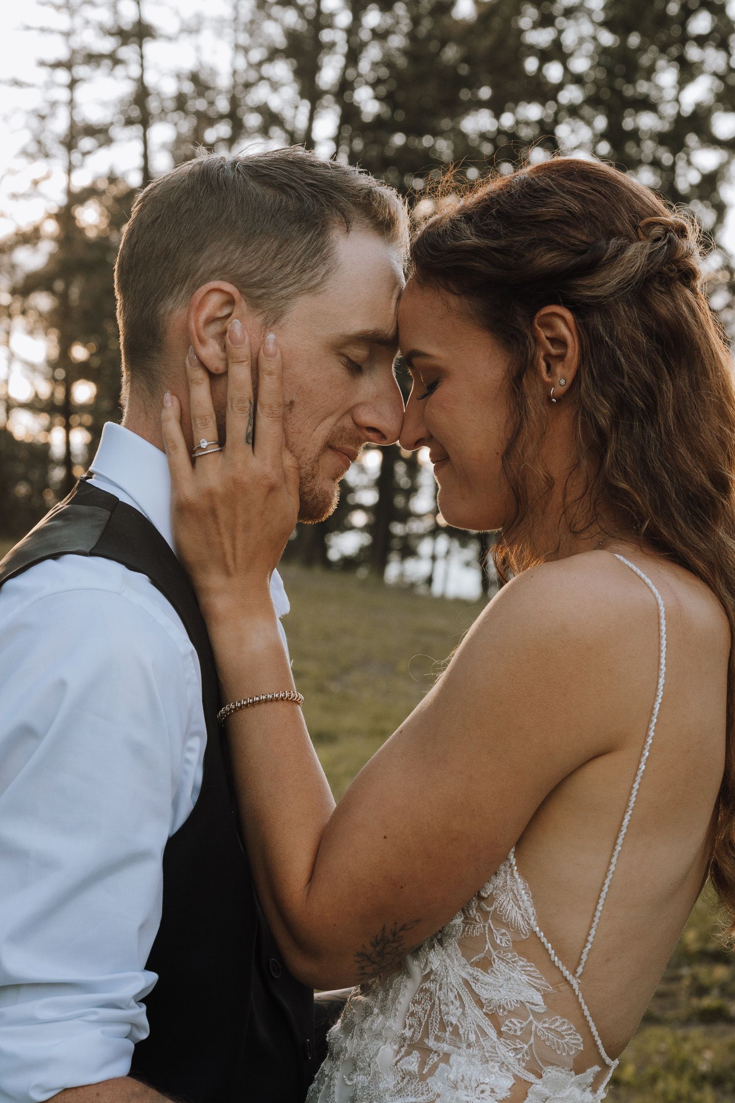A bride and groom share an intimate forehead touch moment outdoors, with the bride in a lace dress and groom in a vest.