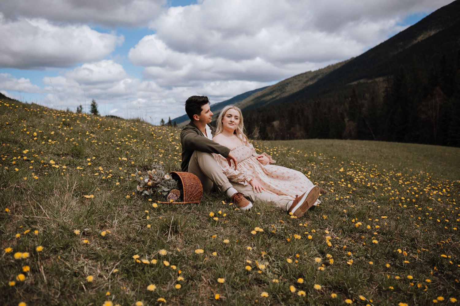 A couple sits together in a wildflower meadow on a mountain slope under dramatic cloudy skies.