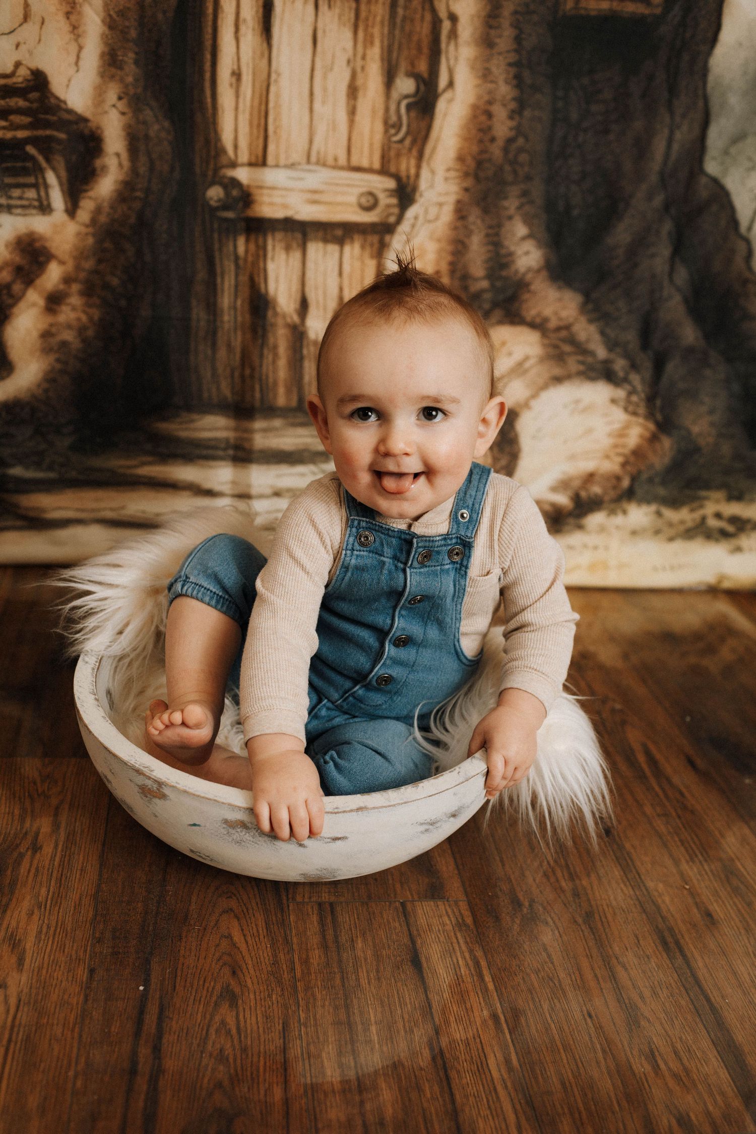 Happy baby in denim overalls sitting in a white bowl against a rustic wooden backdrop.