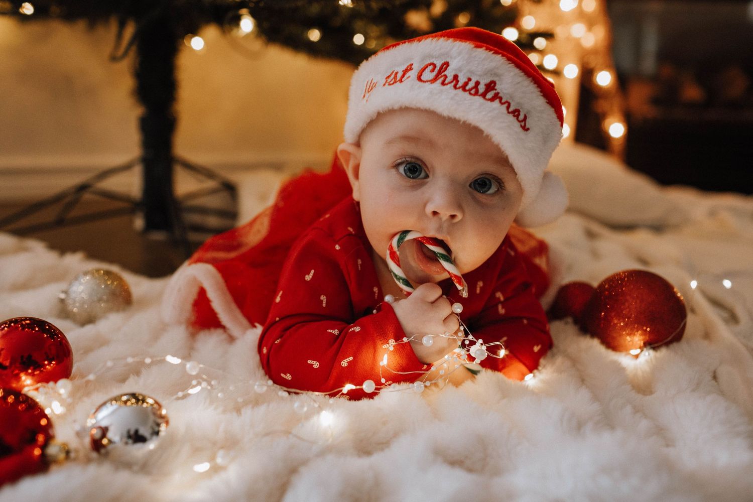 Holiday photo of baby in red outfit lying on white fabric with Christmas ornaments.