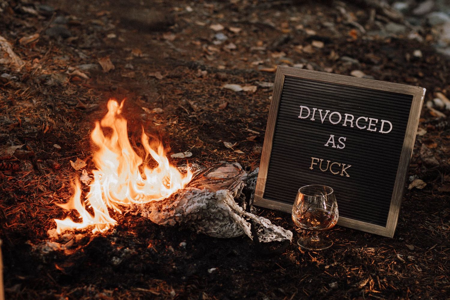 A small campfire burns next to a chalkboard sign reading 'DIVORCED AS F*CK' and a glass of whiskey in the woods.