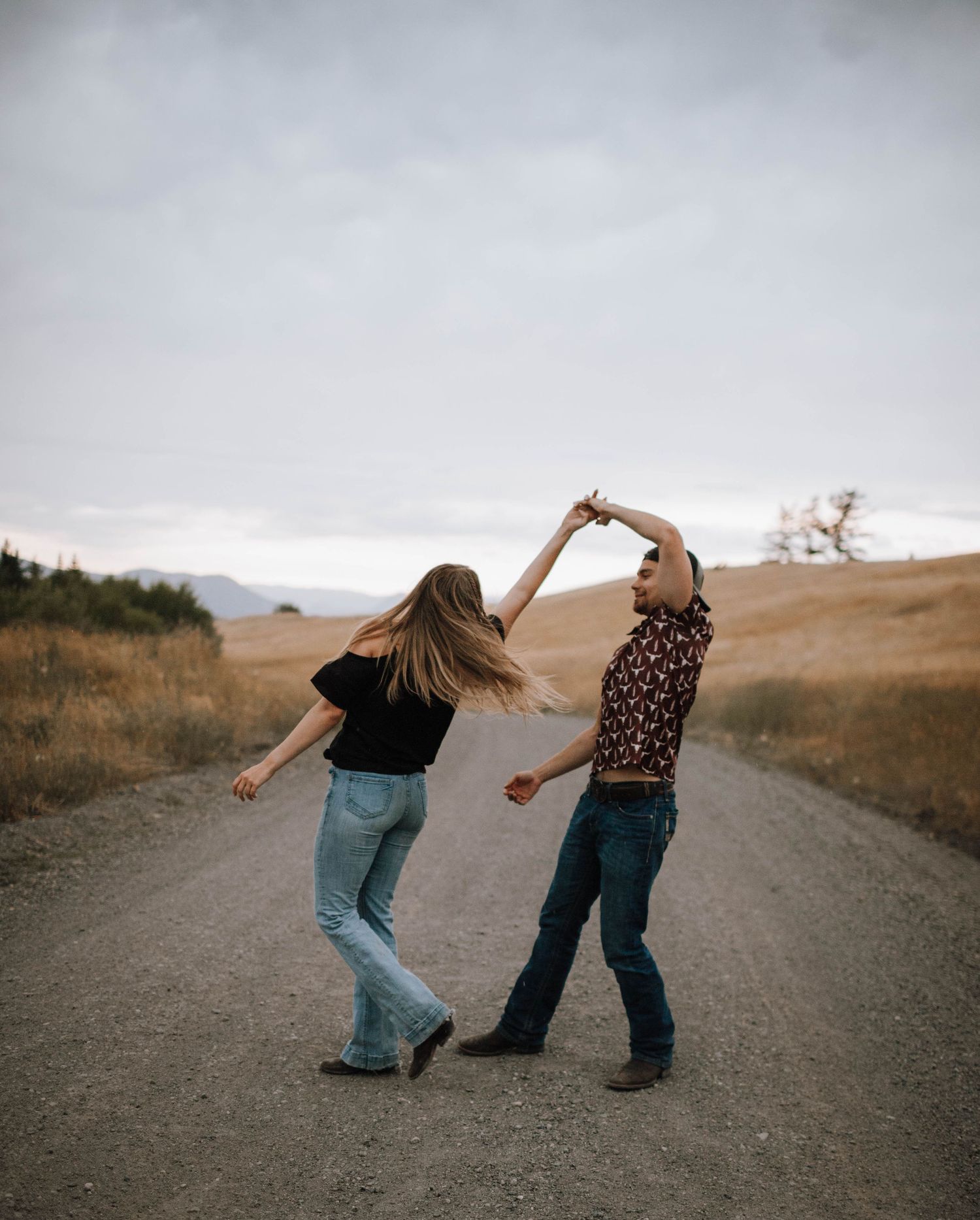 A couple dances playfully on a country road with golden hills in the background.