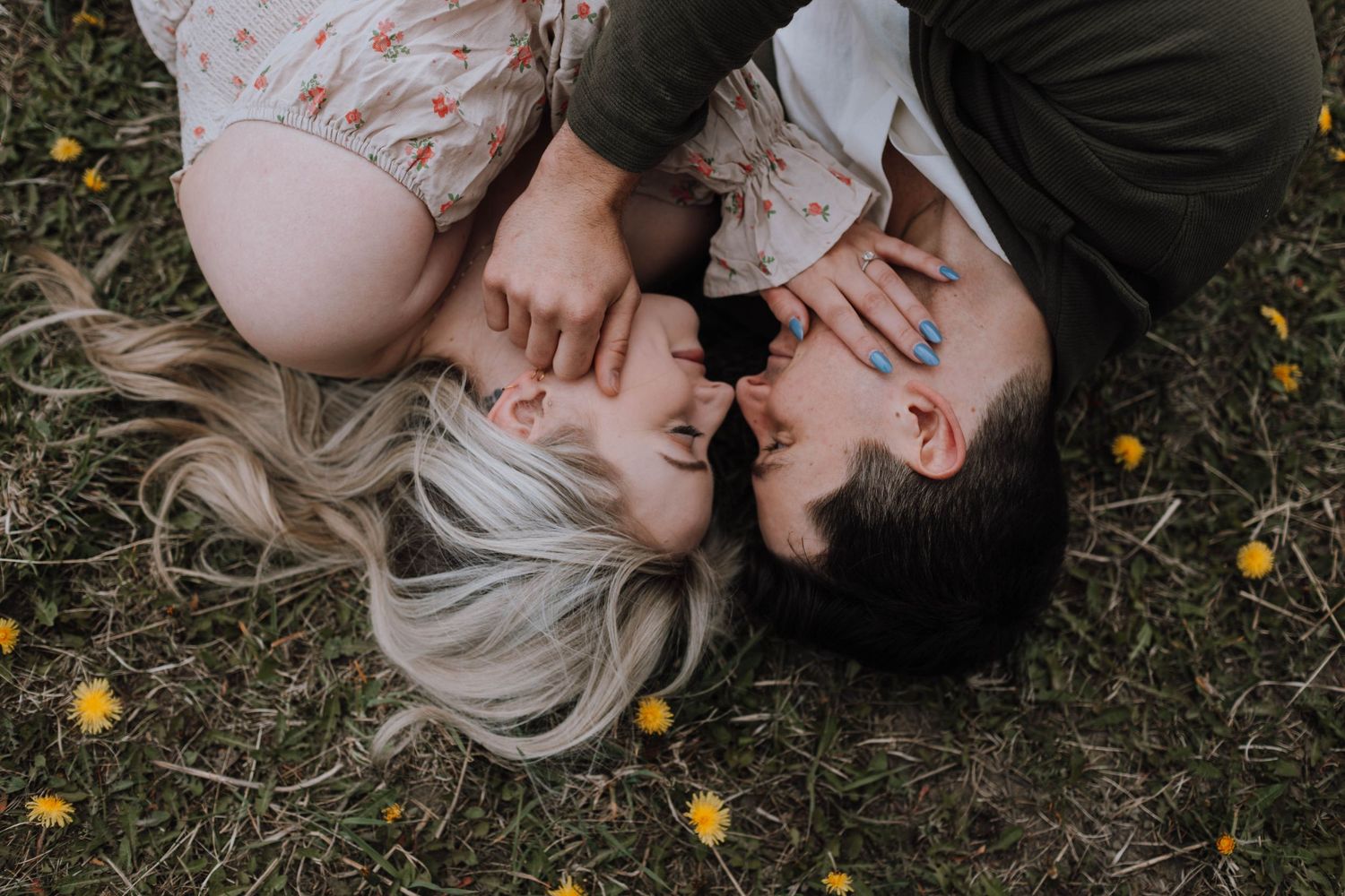 Romantic couple lying in grass with scattered yellow dandelions during an intimate outdoor photoshoot.