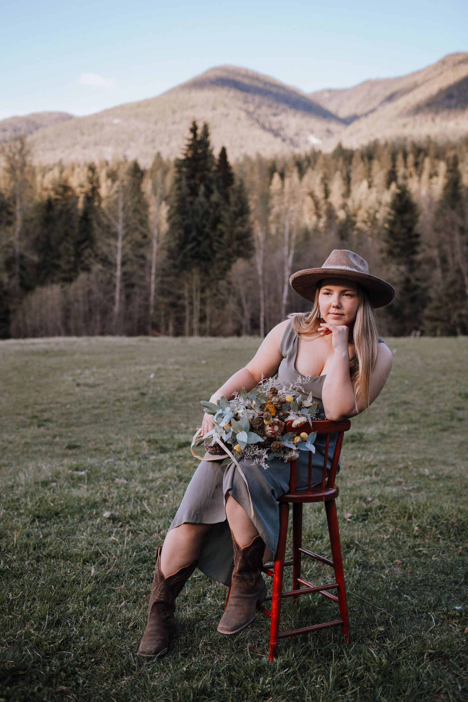 Person in hat sitting on red stool with mountain landscape and evergreen forest in background.