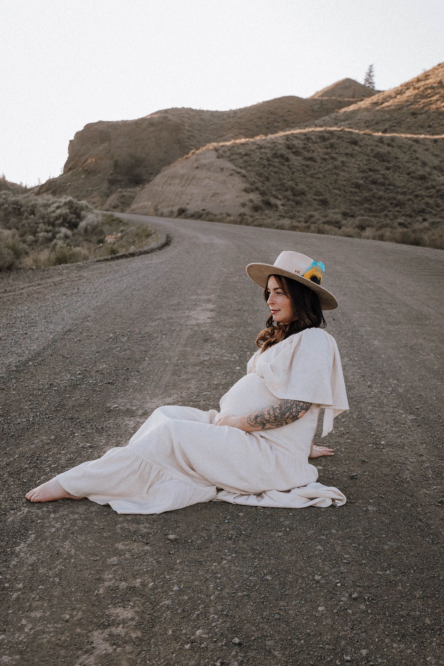 A person in a flowing white dress and brown hat reclines on a desert road at sunset.