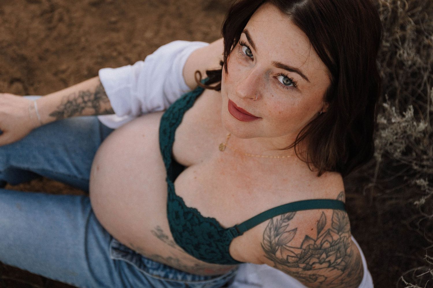 Person in teal bralette and jeans lounging against stone wall, showing arm tattoos in outdoor portrait.
