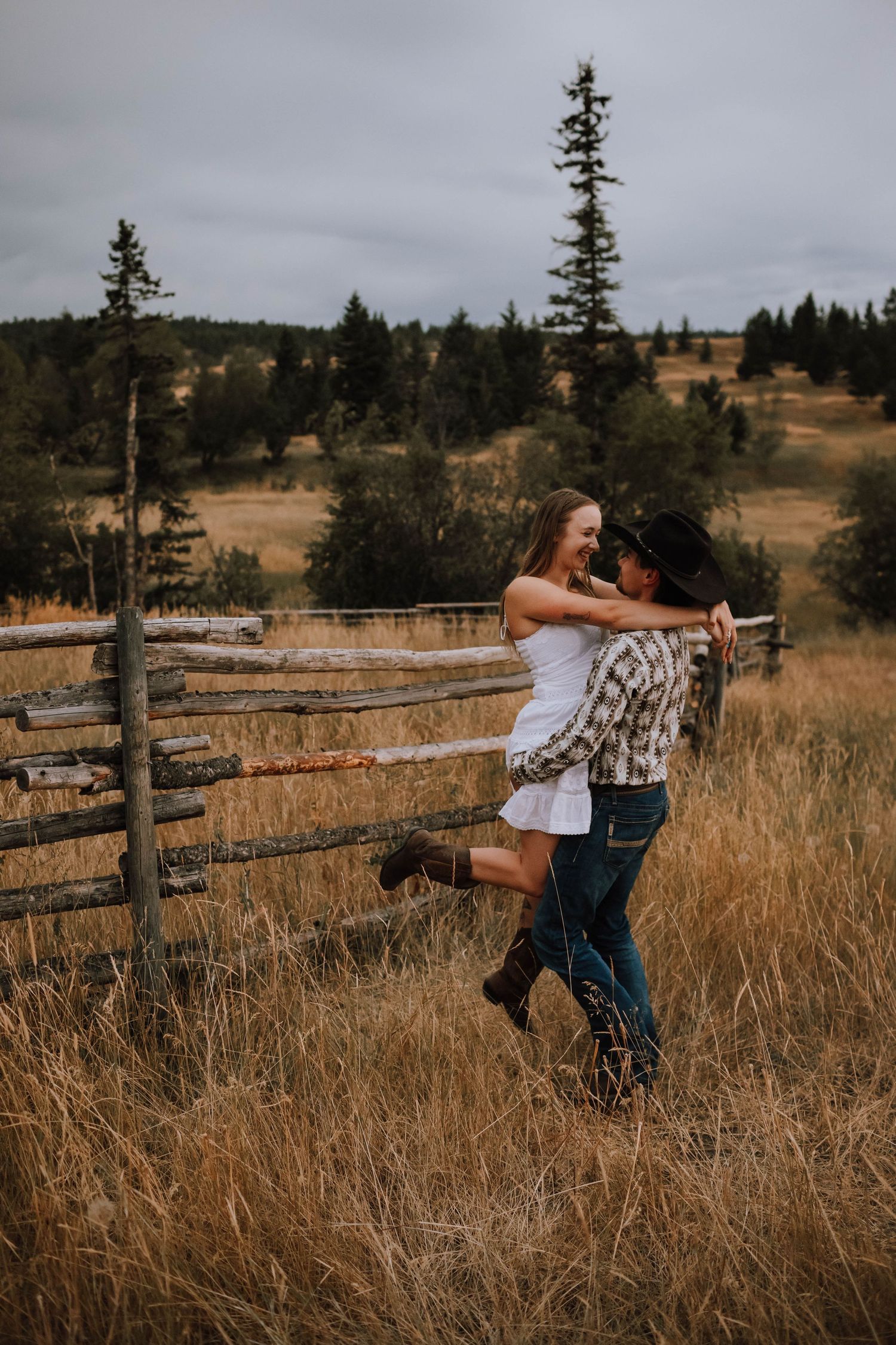 A couple embraces by a wooden fence in a scenic mountain meadow at golden hour.