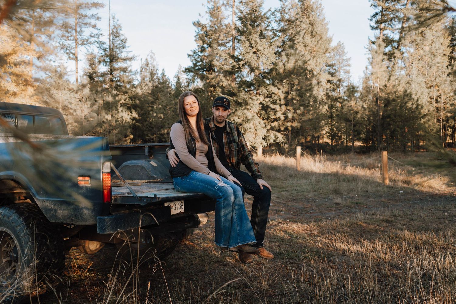 A couple sits on the tailgate of their blue GMC pickup truck in a forest clearing.
