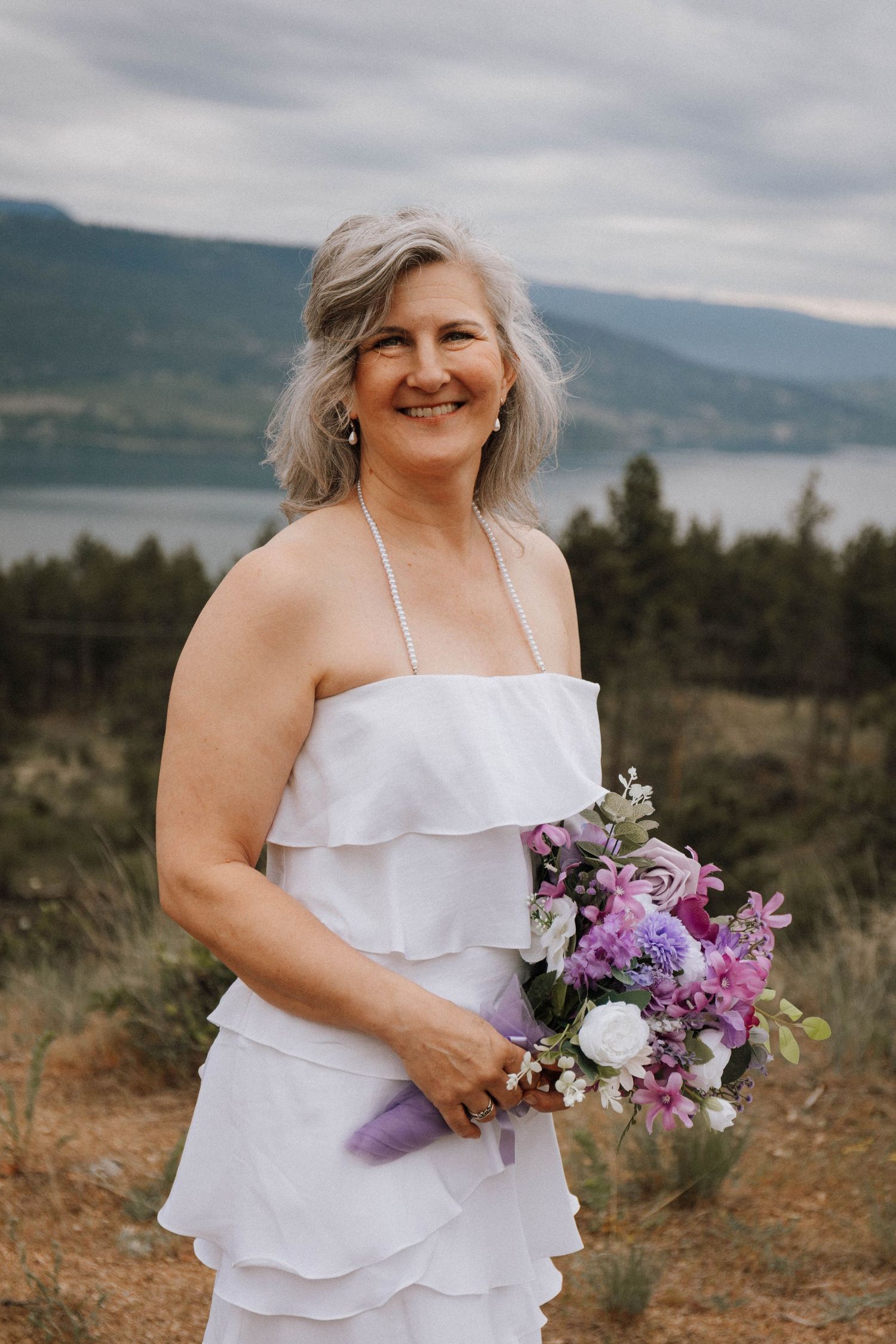 Person in white tiered halter dress holding vibrant purple and pink flower bouquet against mountain lake landscape backdrop.