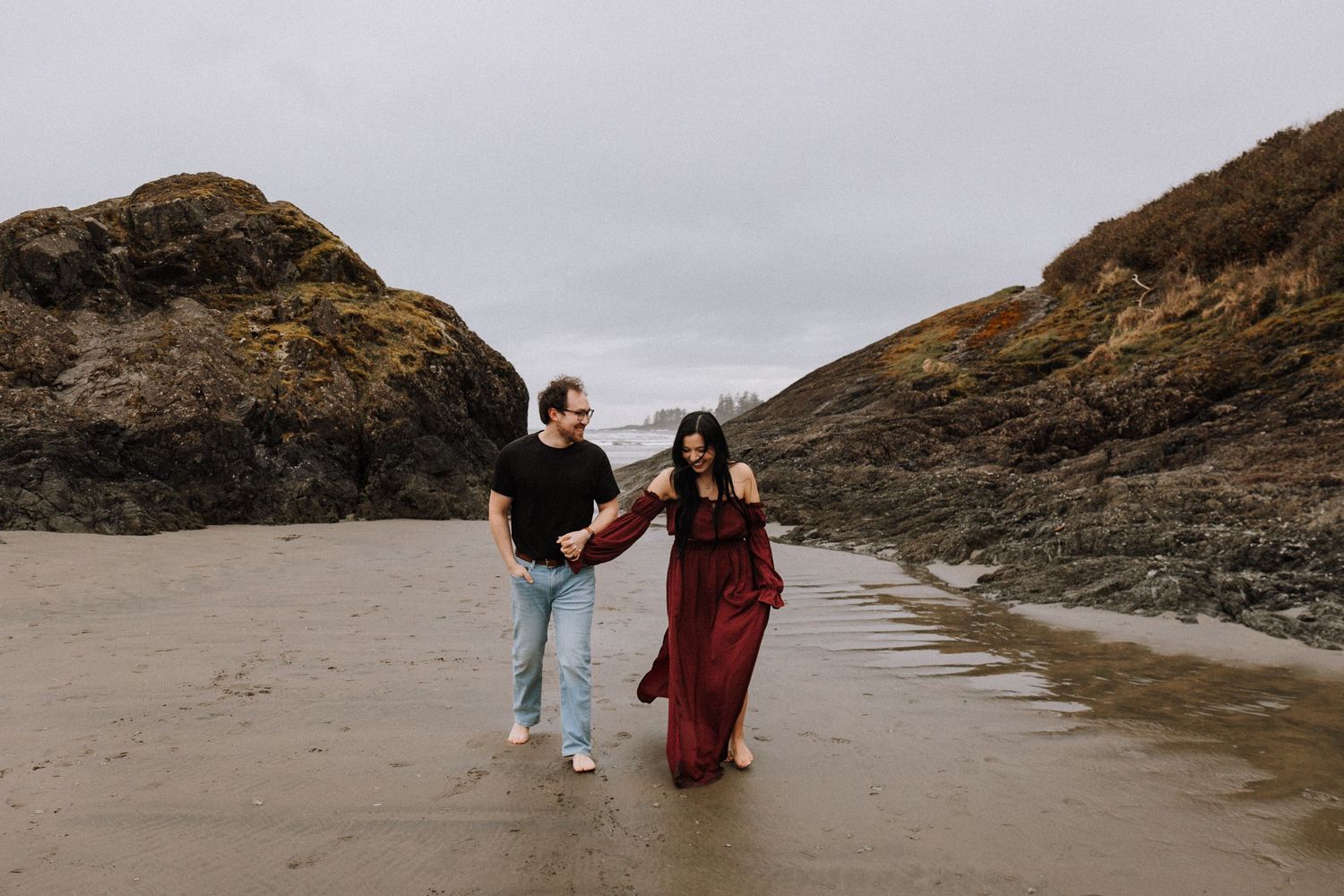 Couple walking hand in hand on a rocky beach, woman in red dress, man in black shirt and jeans, overcast sky. Long Beach Tofino BC