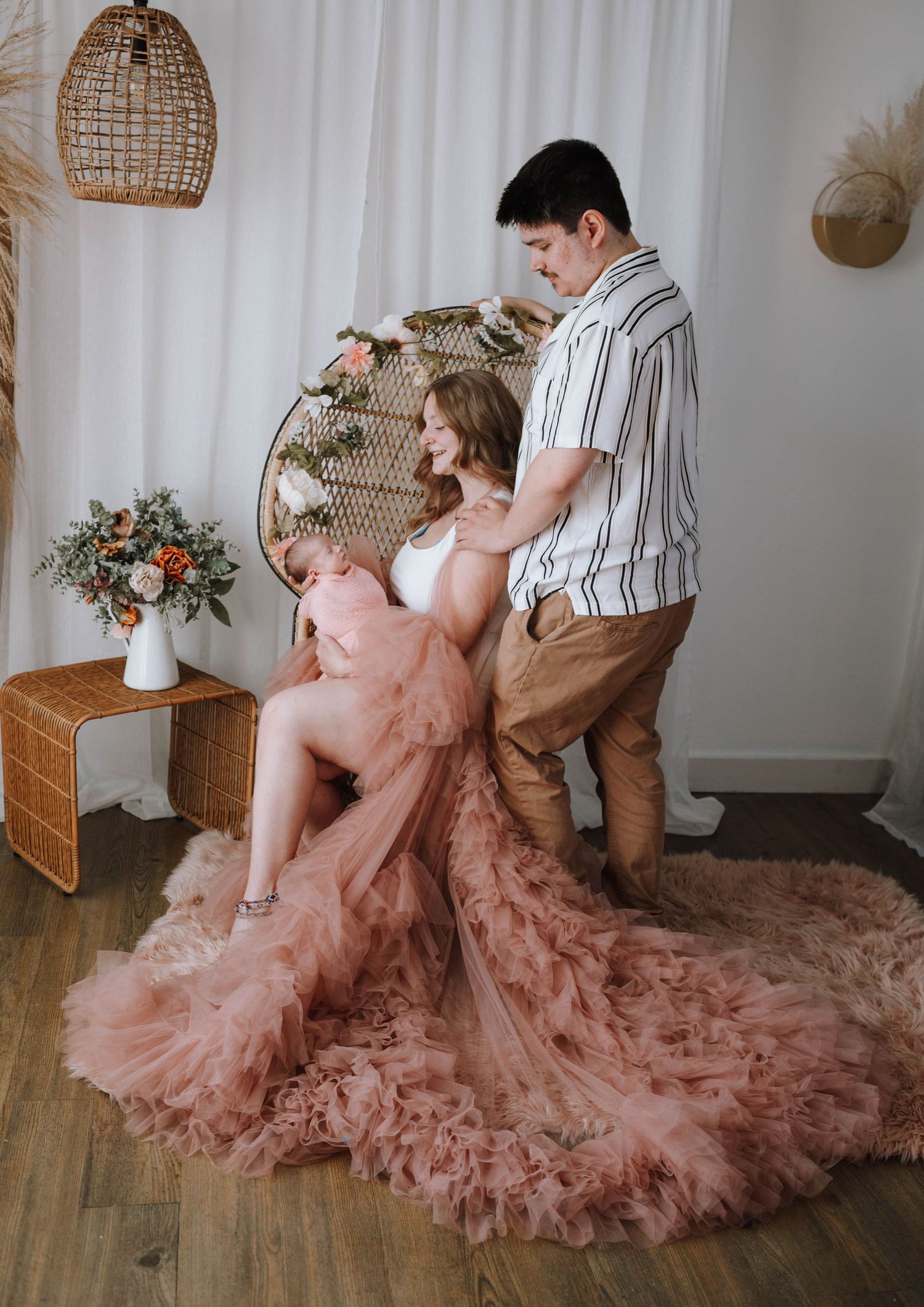 A romantic maternity photo featuring a flowing pink tulle dress and rattan decor in a bohemian styled indoor setting.