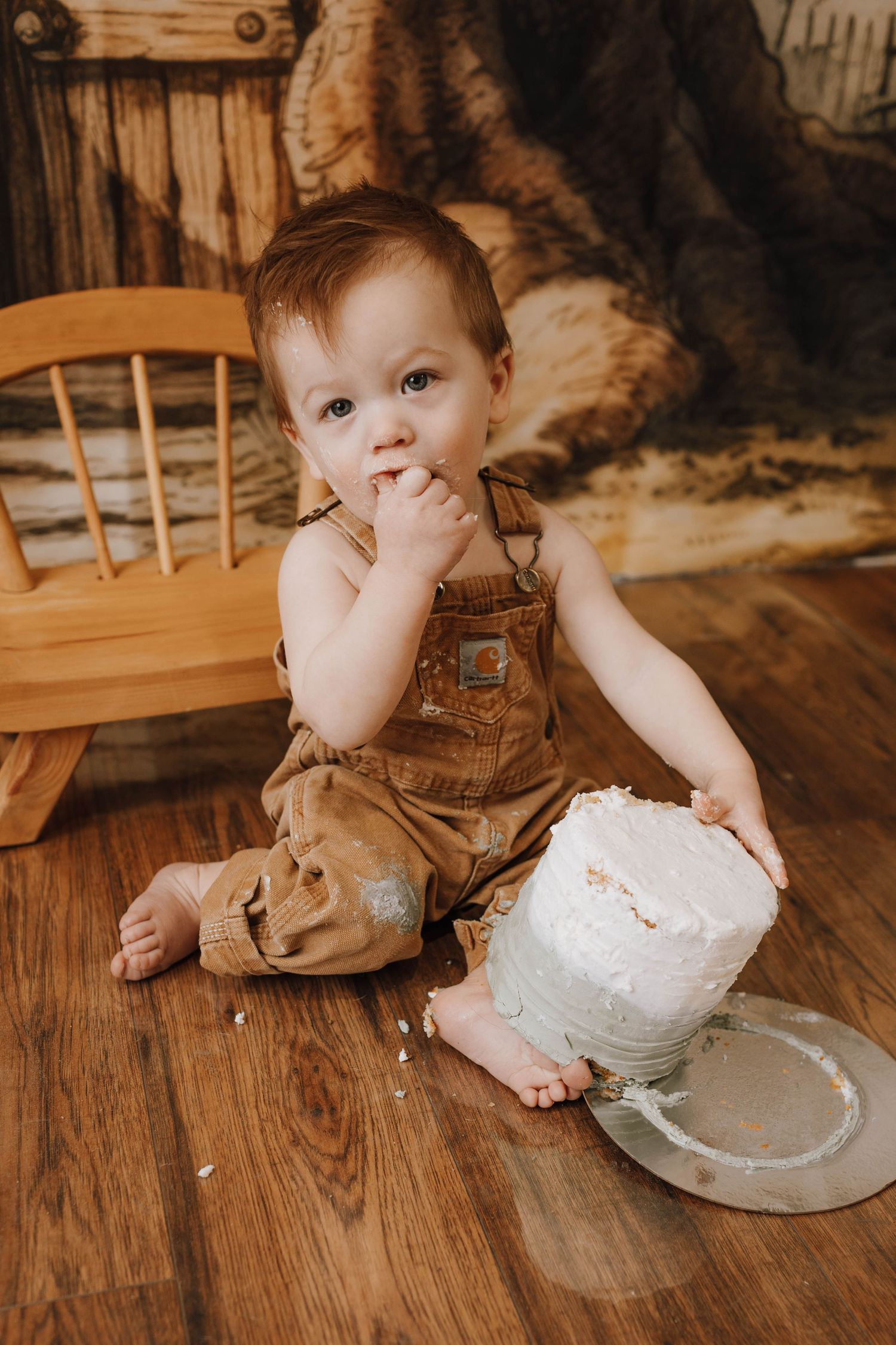 Baby in brown overalls sitting beside white birthday cake on hardwood floor.