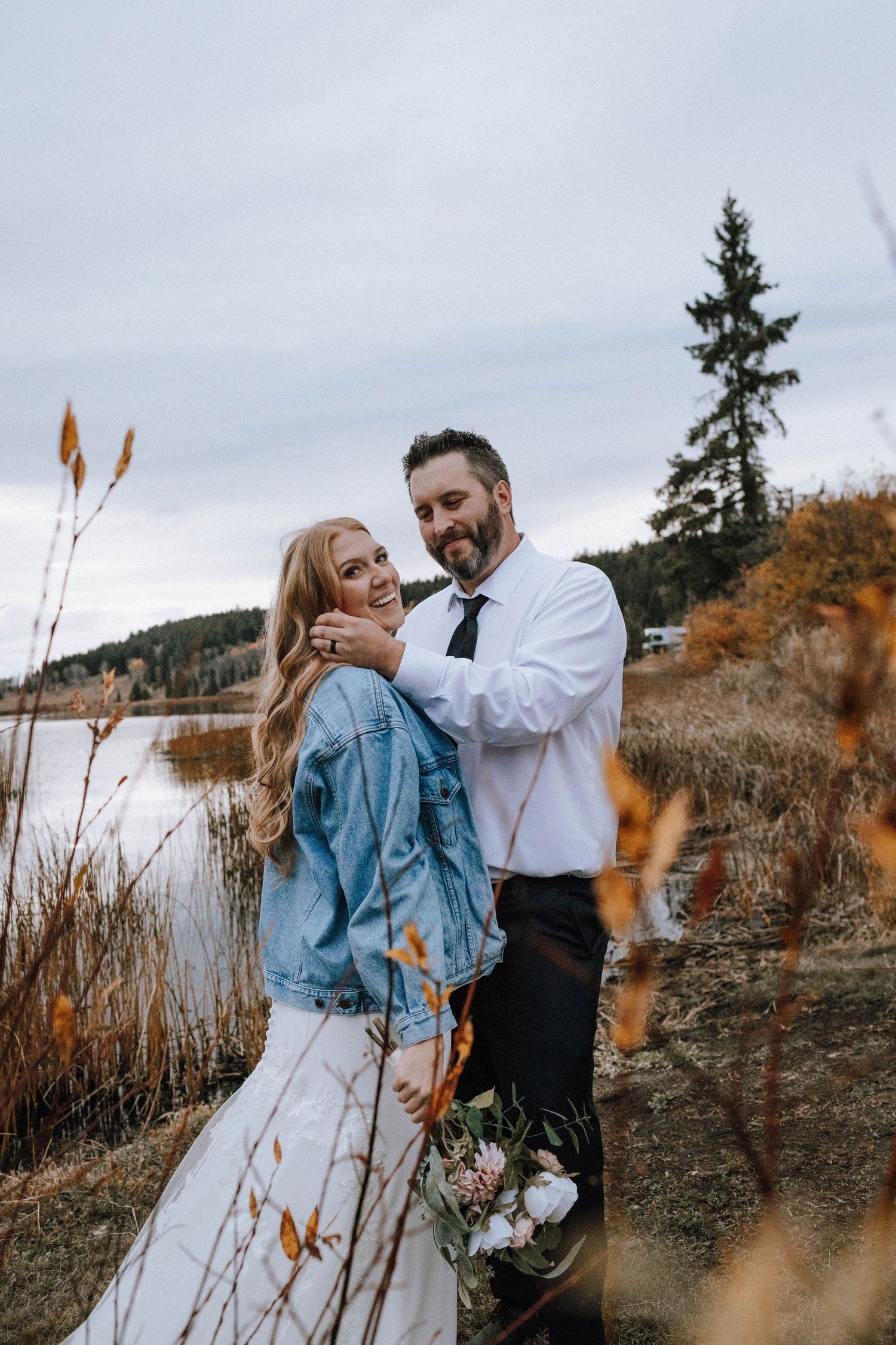 Couple embraces by a lake during autumn with tall grasses and mountains in background.