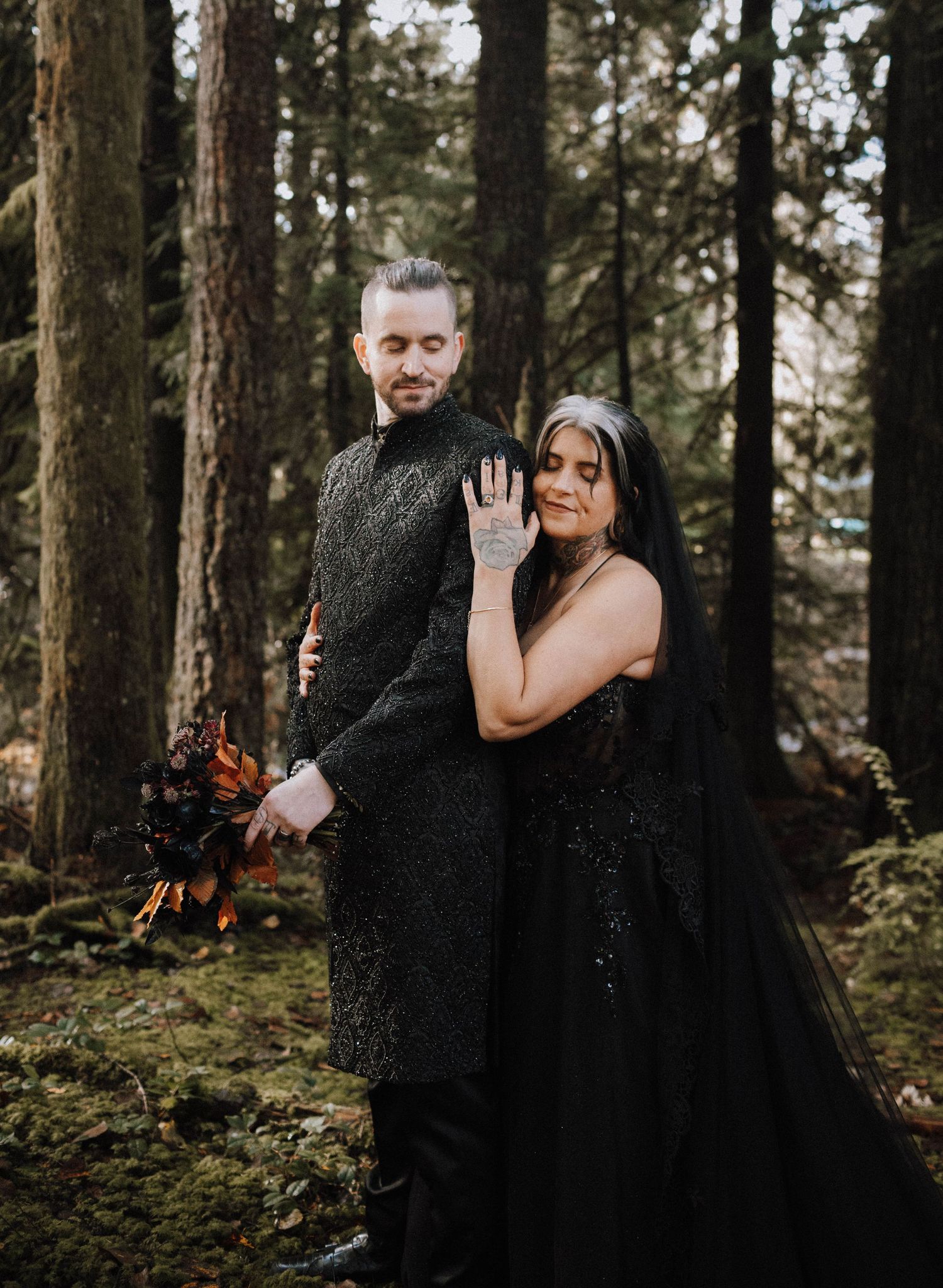 A couple in matching black formal wear holds hands in a misty evergreen forest.