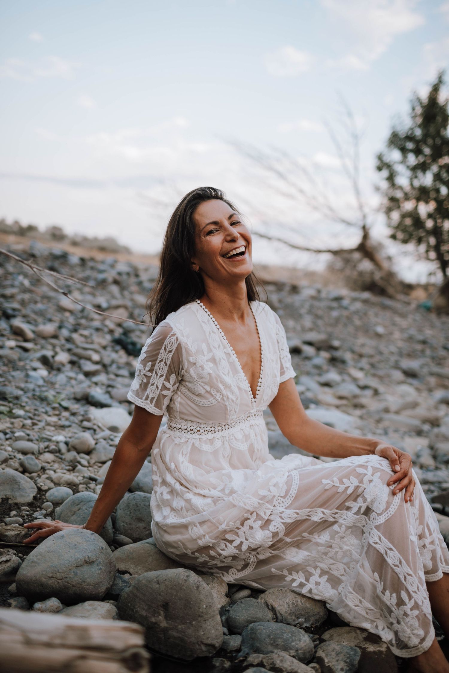 Person in white lace dress laughing while sitting on rocky terrain at sunset.