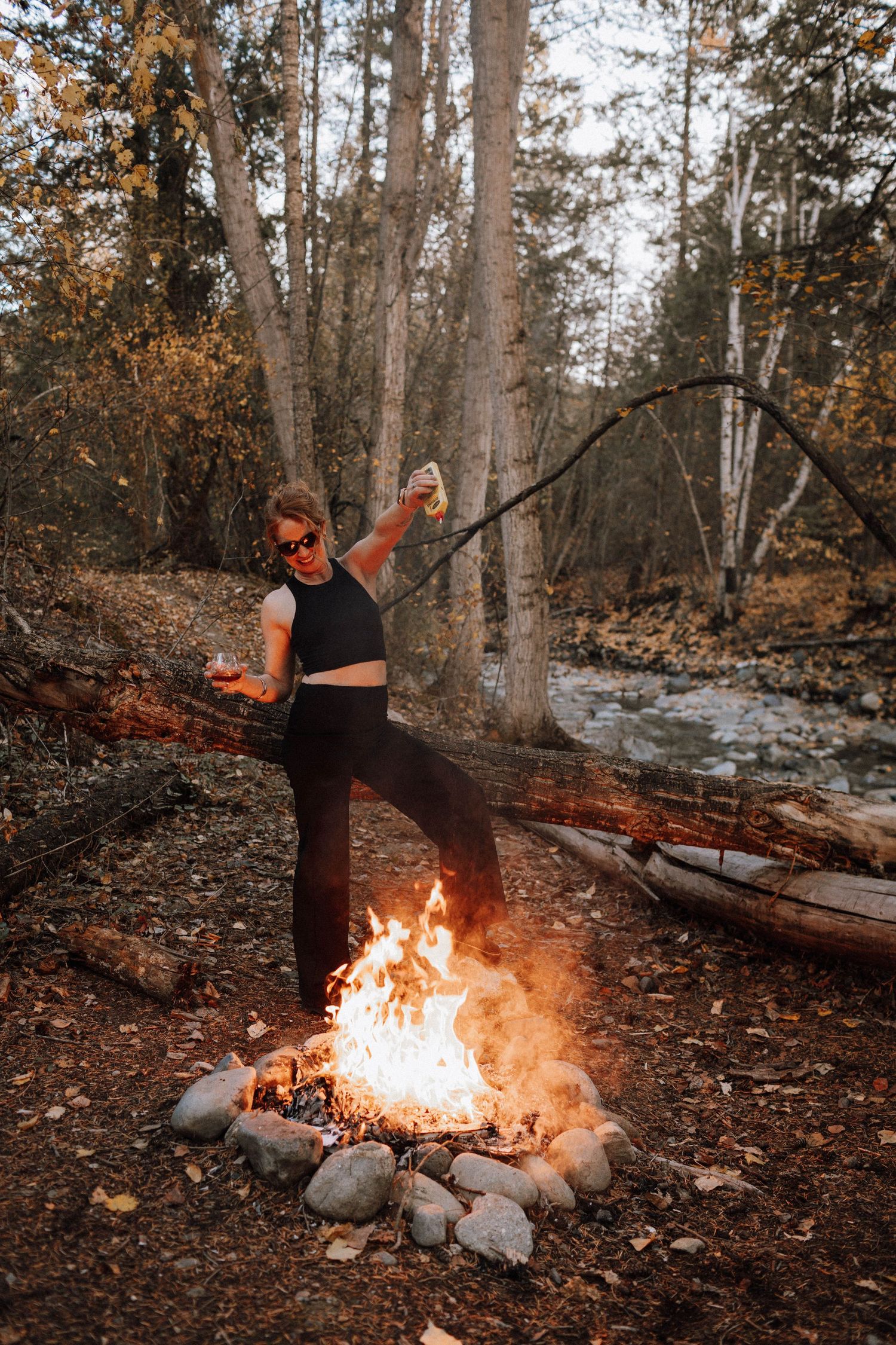 A person in dark clothing stands near a bright campfire surrounded by autumn leaves and birch trees in a forest setting.