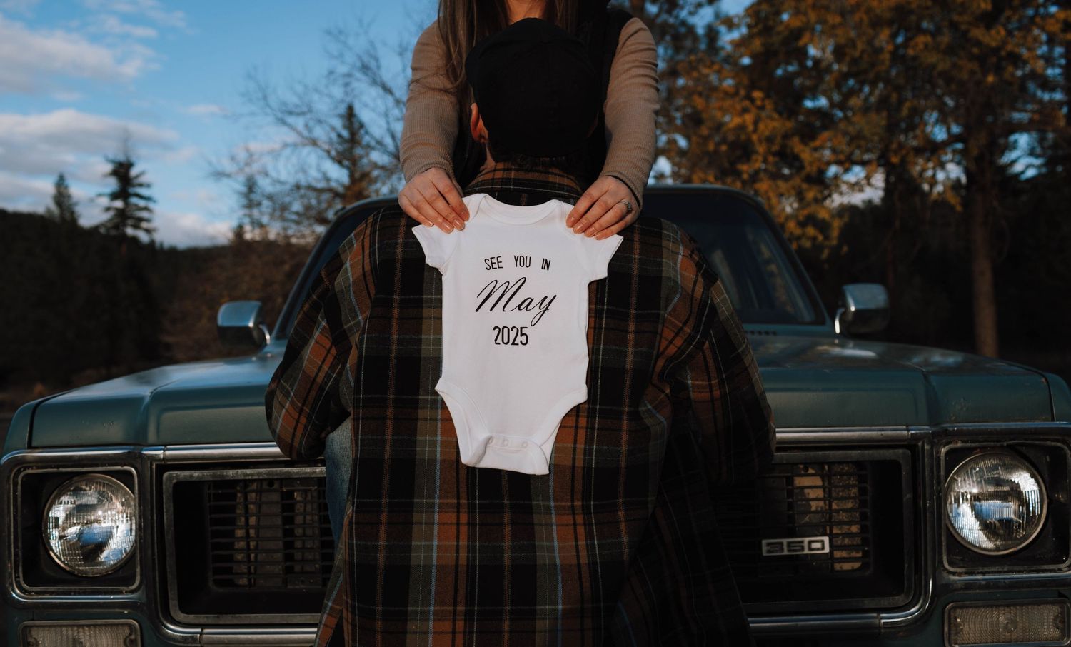 A white baby onesie is held up against vintage trucks in an autumn setting.
