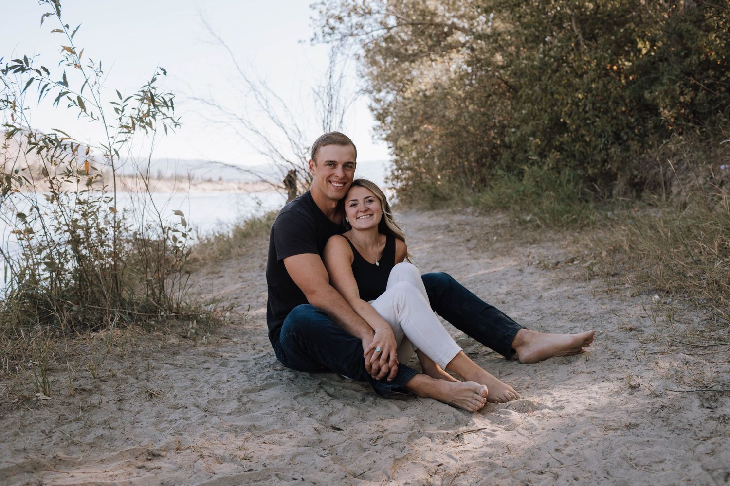 A casual couple sits together on a sandy path near water during an outdoor photoshoot.