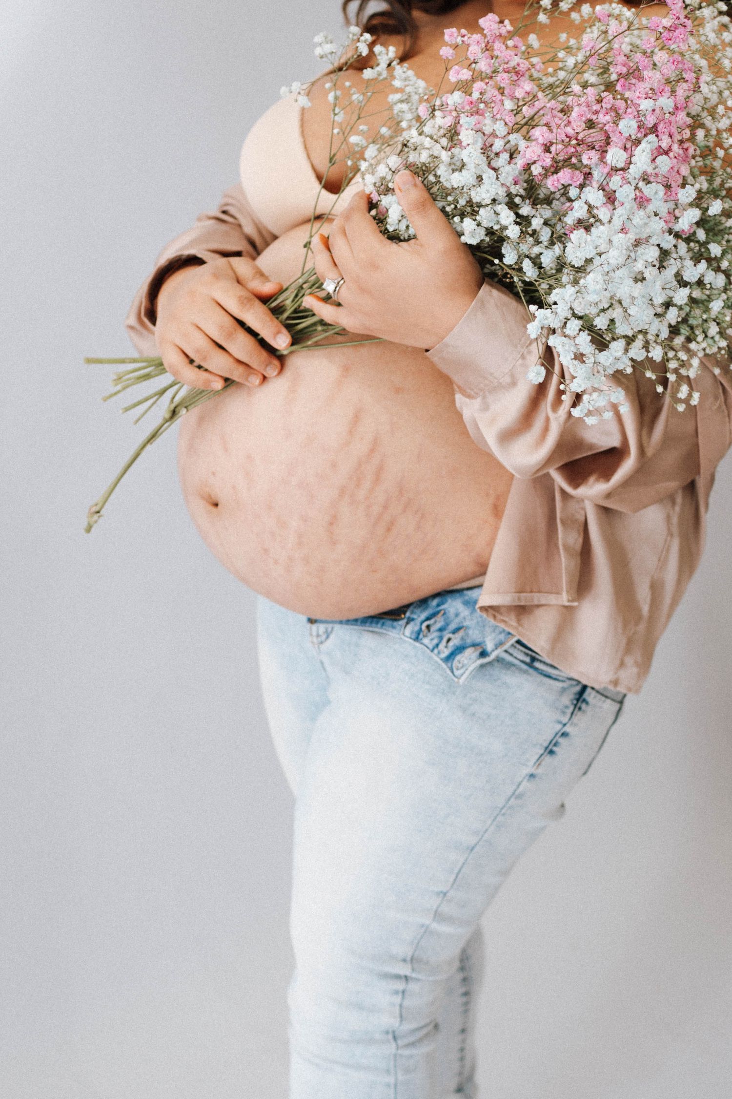 A pregnant belly in light jeans and peach blouse holding delicate baby's breath and pink flowers against grey background.