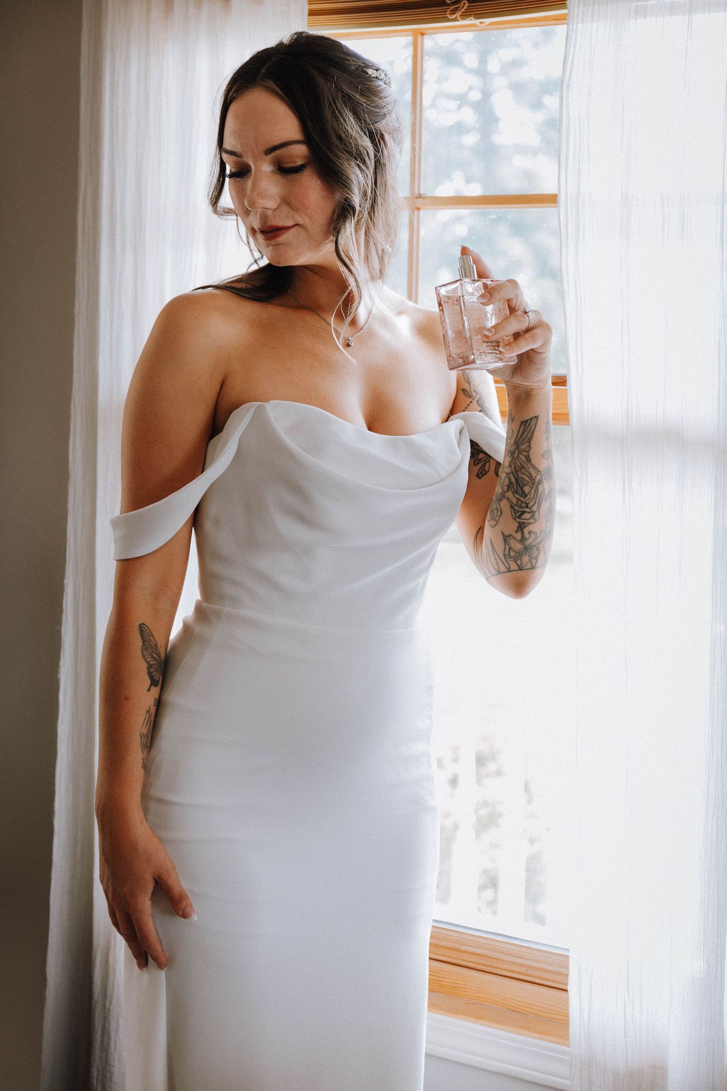 Person in white off-shoulder dress stands by window in soft natural light, holding a glass.