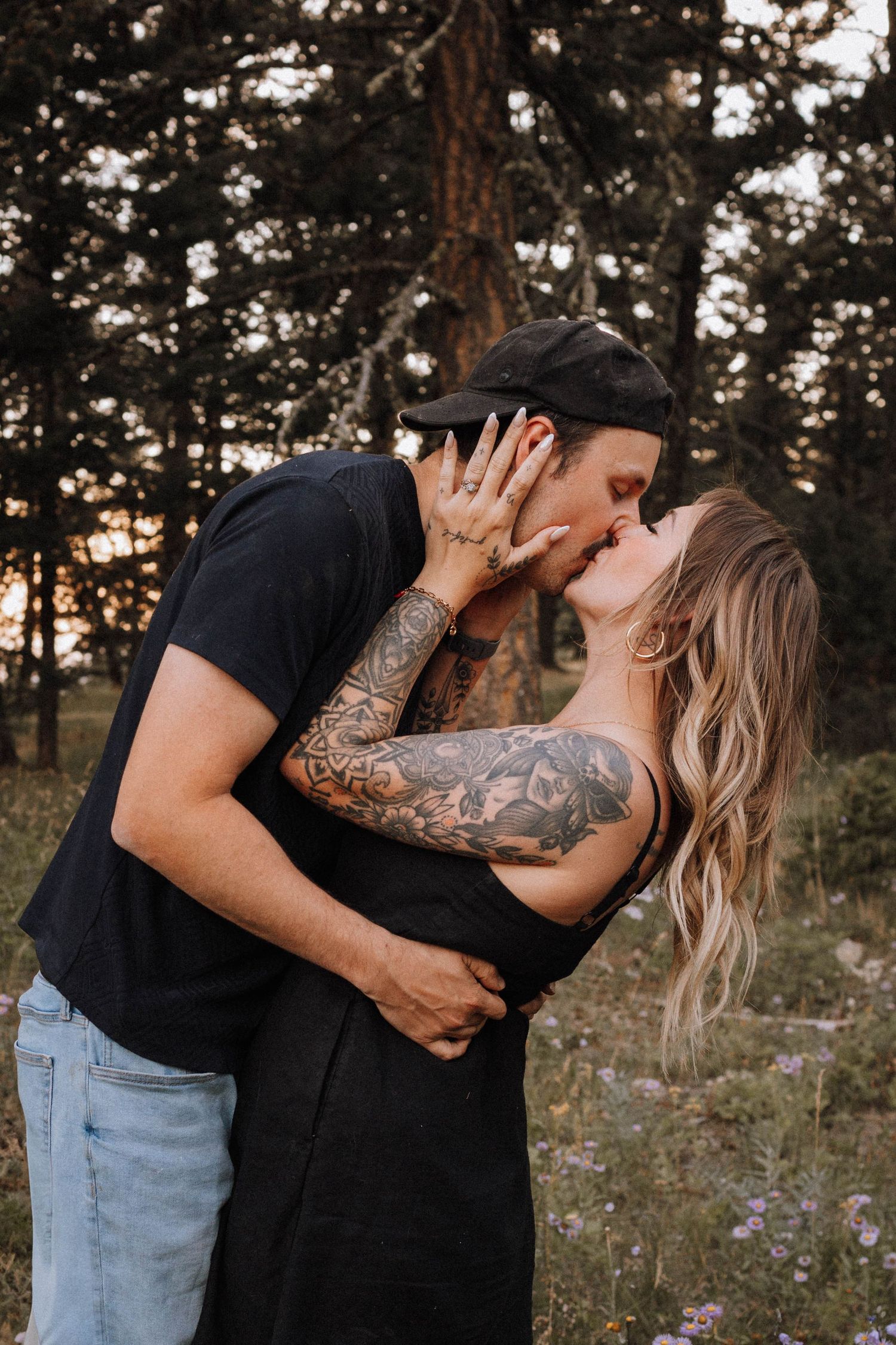 A couple embraces in a romantic outdoor photoshoot with wildflowers and trees in the background at sunset.