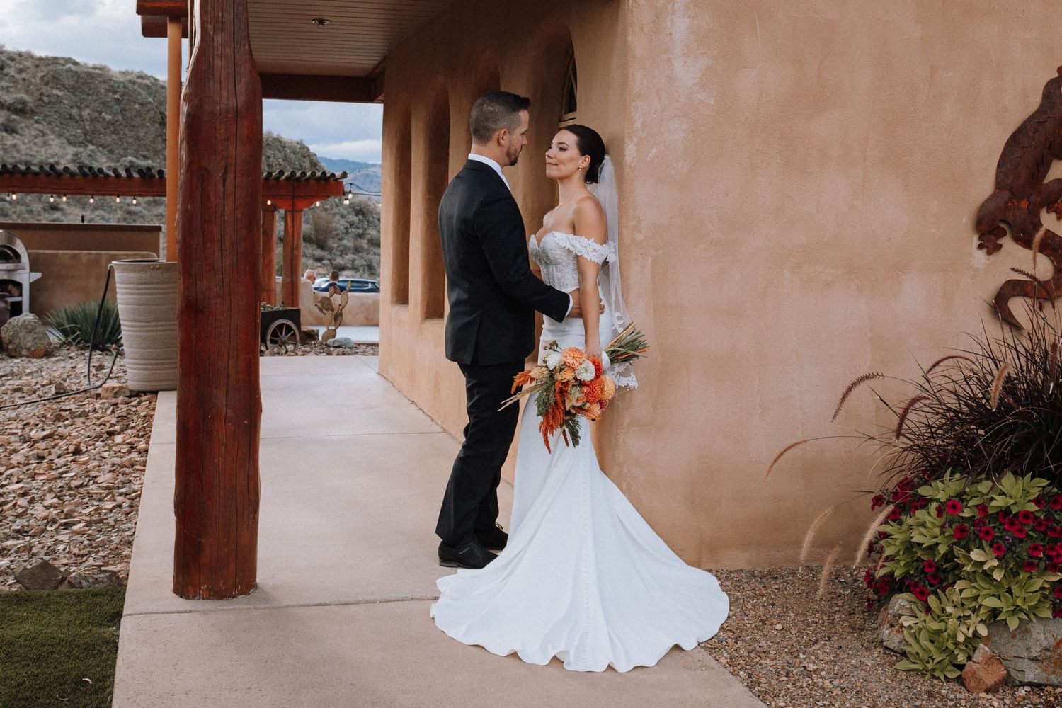 Couple in wedding attire standing under adobe archway with orange floral bouquet.